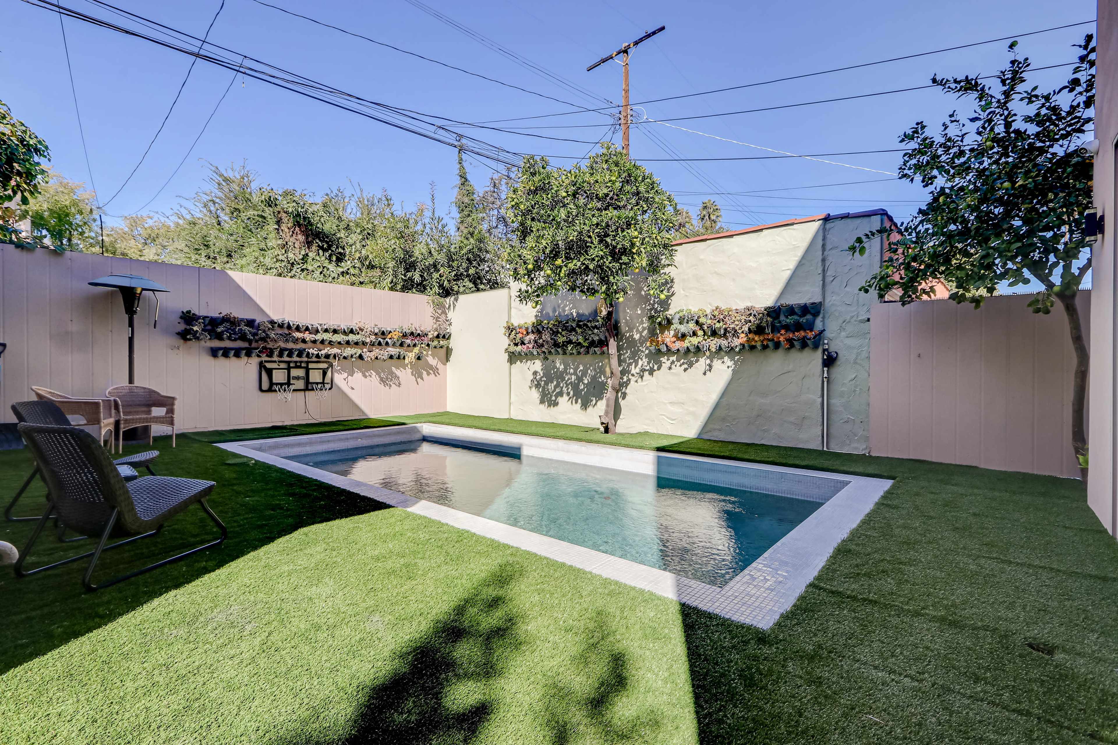 The image shows a small, rectangular swimming pool surrounded by artificial turf and bordered by walls with greenery and outdoor seating.