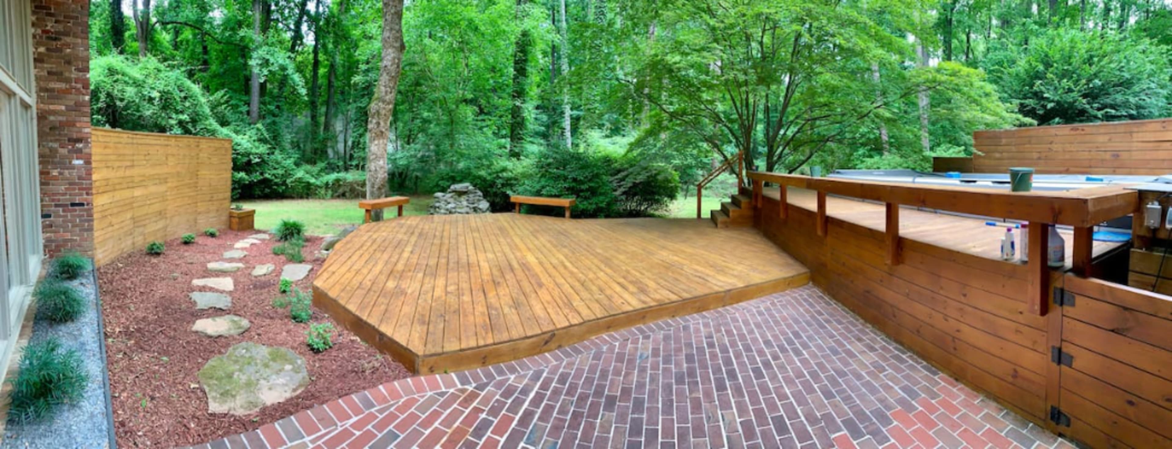 The image shows a wooden deck surrounded by greenery, with a brick pathway leading to a landscaped area featuring rocks and plant beds.