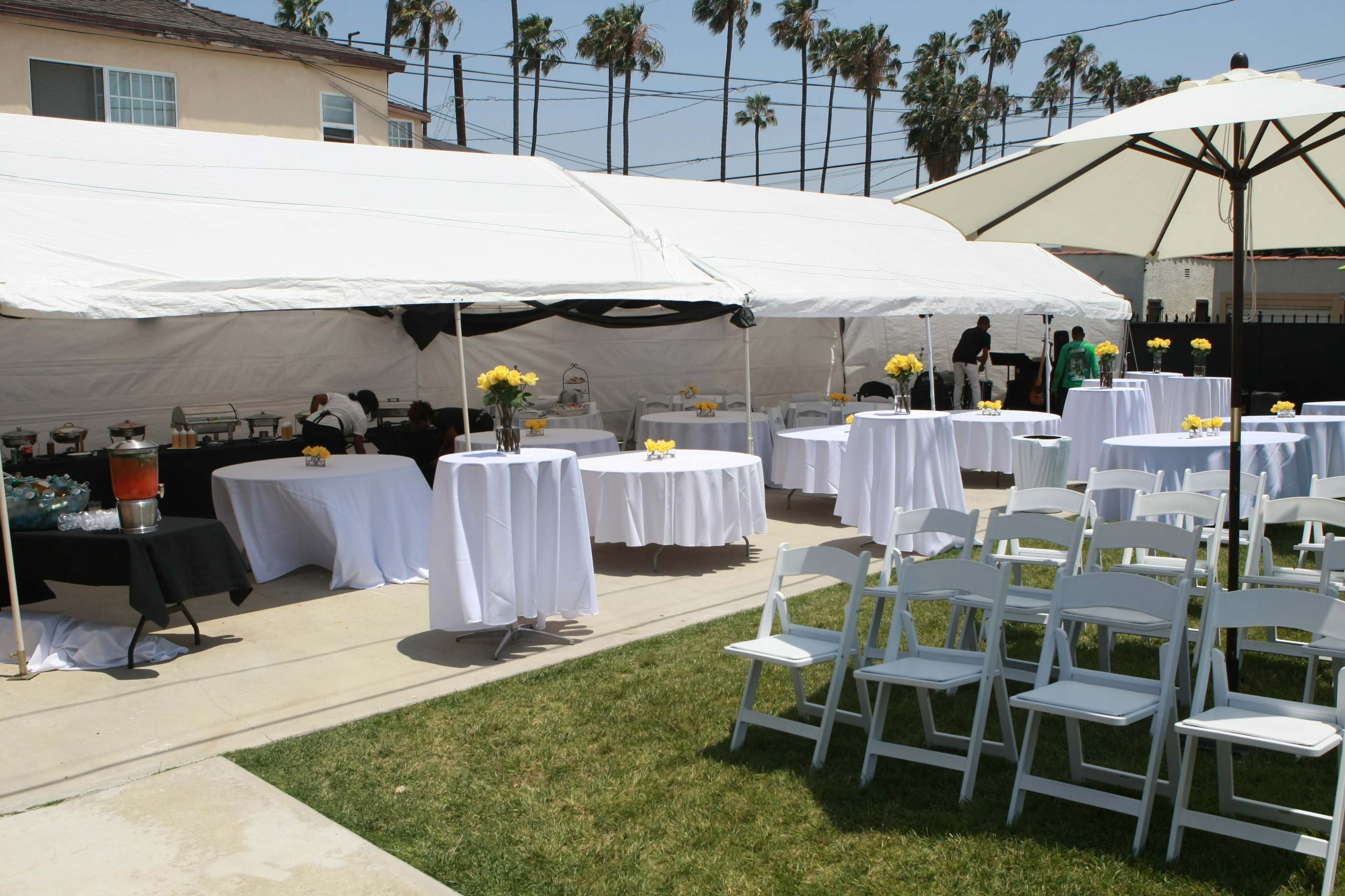 A tented outdoor area is set up for an event, featuring round tables with white tablecloths, chairs, and decorations, surrounded by palm trees.