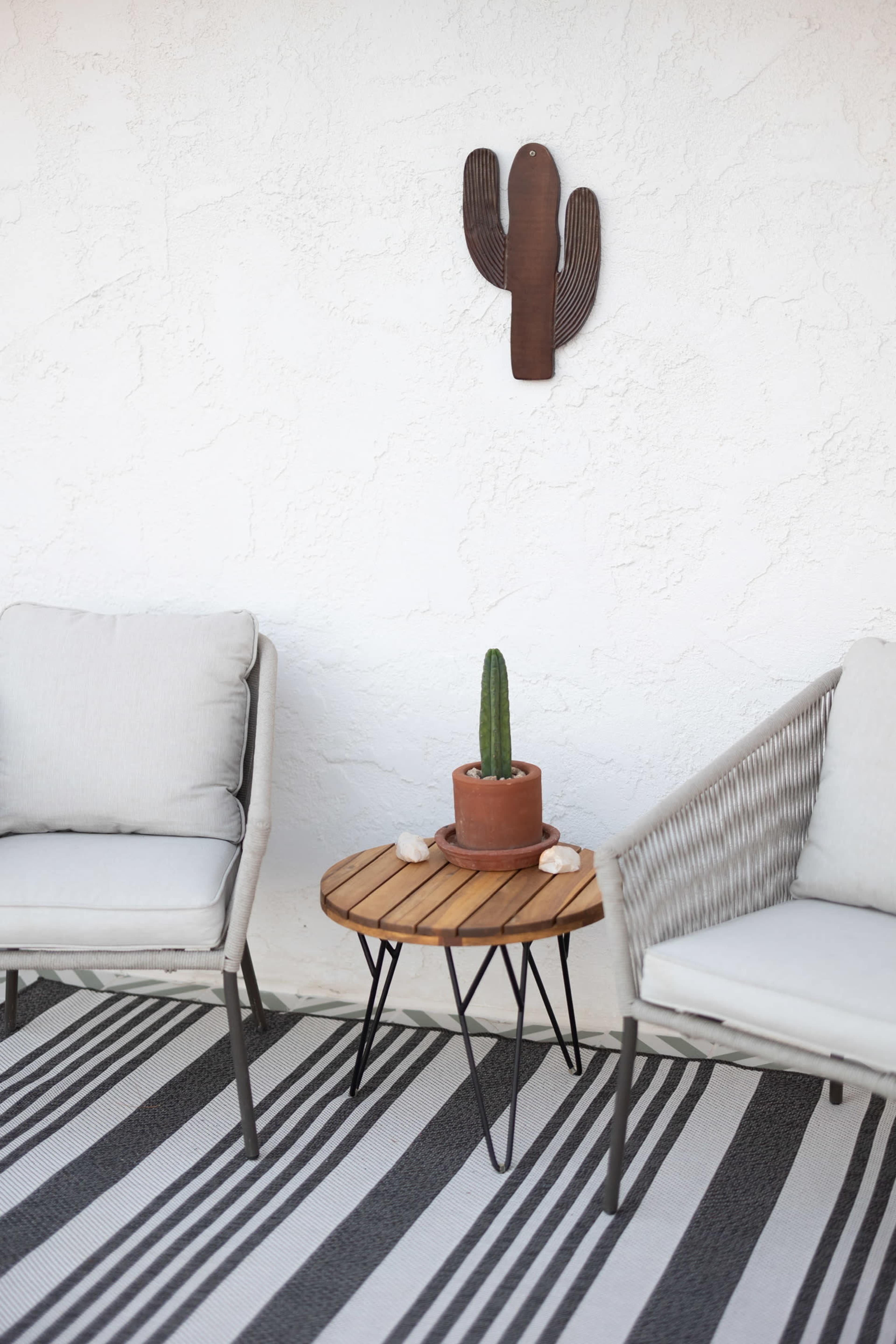 The image shows a small outdoor seating area with two chairs, a round wooden table with a potted cactus, and a striped rug on a textured white wall backdrop.