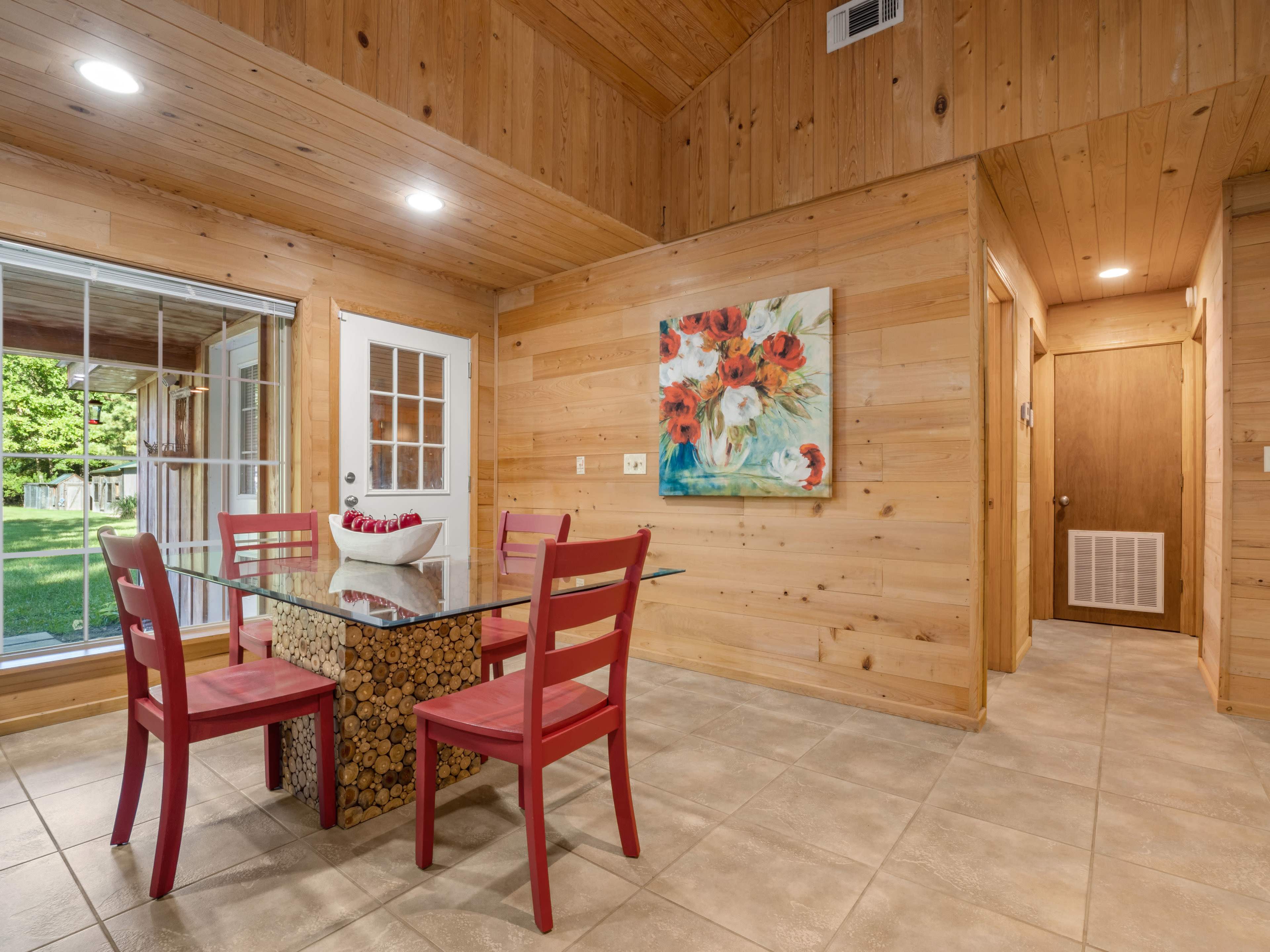 A dining area with a wooden table and red chairs, featuring a large window and wood-paneled walls.
