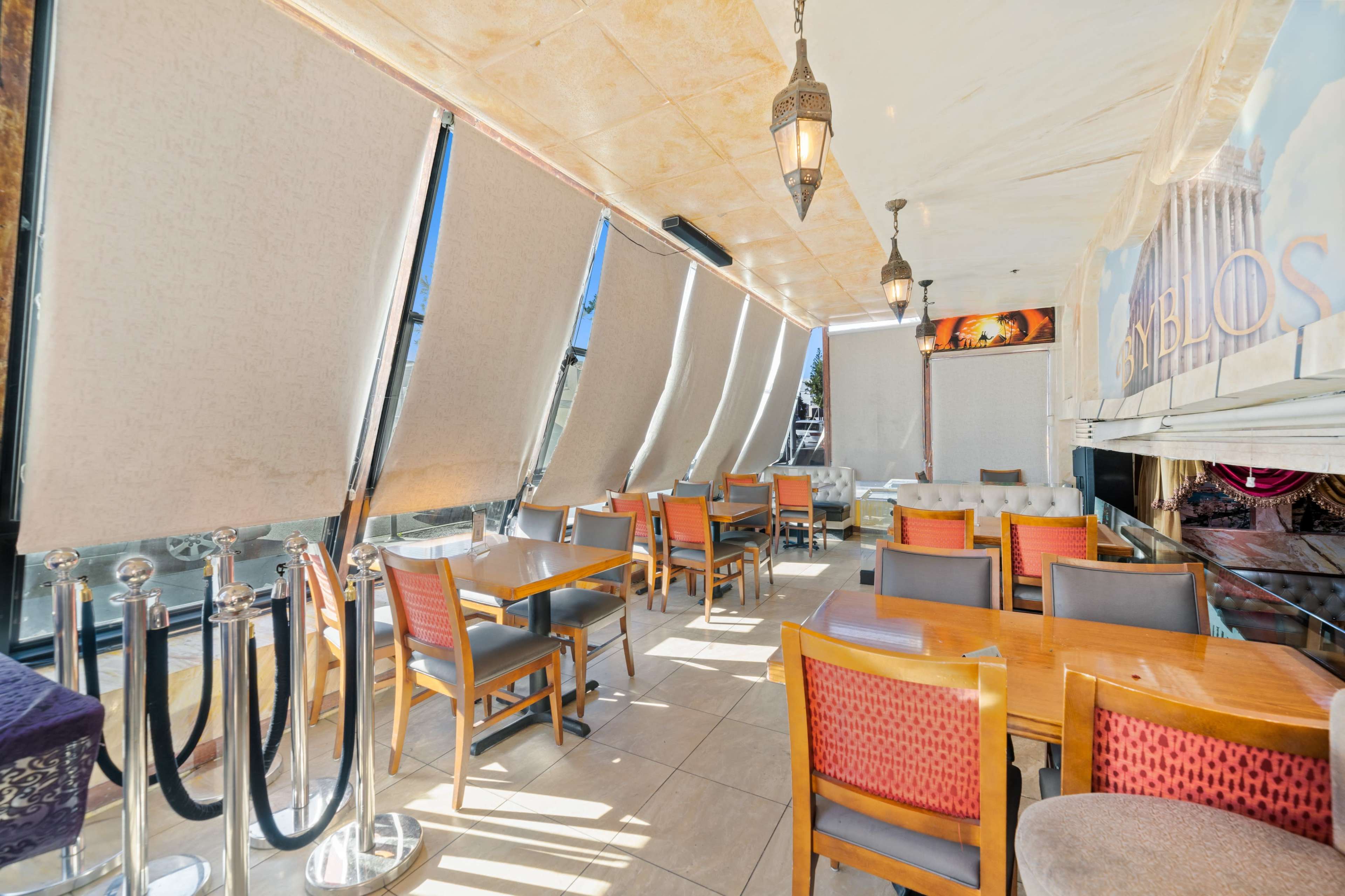 A restaurant interior with large windows covered by fabric shades, wooden tables, and chairs arranged throughout the space.