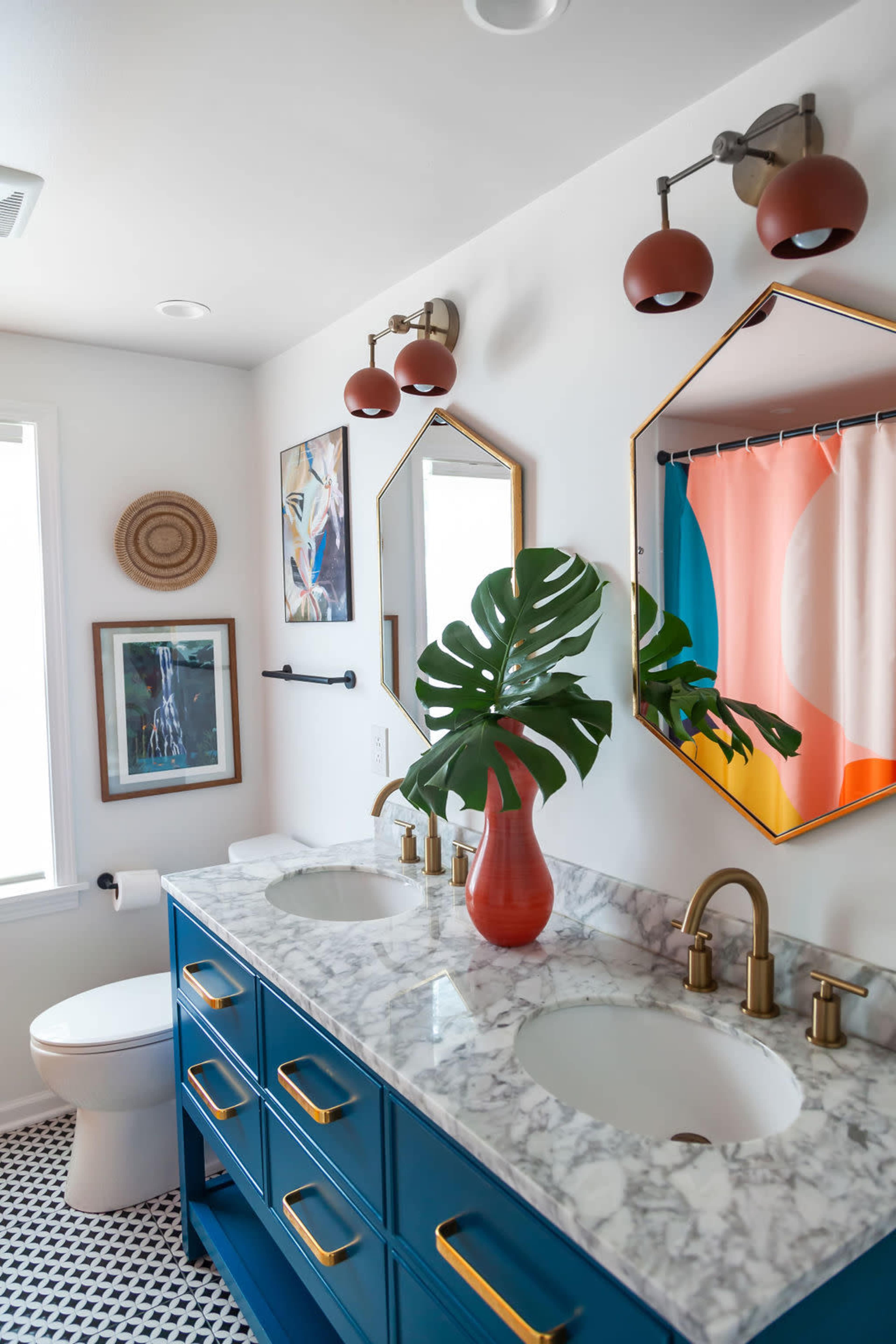 A bright bathroom featuring a double sink vanity with a blue base, marble countertop, decorative lighting, and colorful shower curtain, along with framed artwork and a large green plant as a centerpiece.