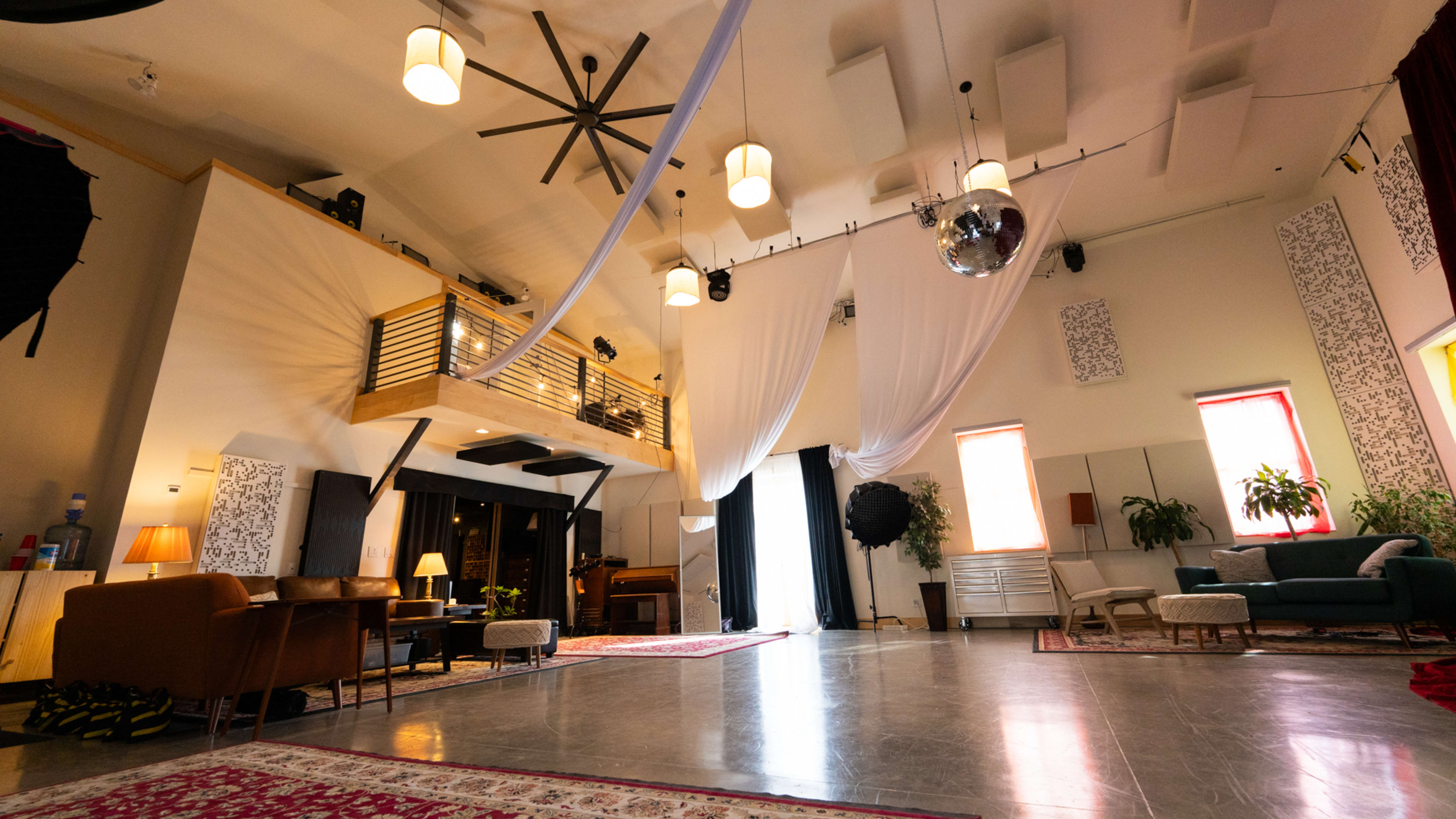 The image features a spacious, well-lit interior with a high ceiling, lounge seating arranged around a large open area, and decorative fabric draped from above.