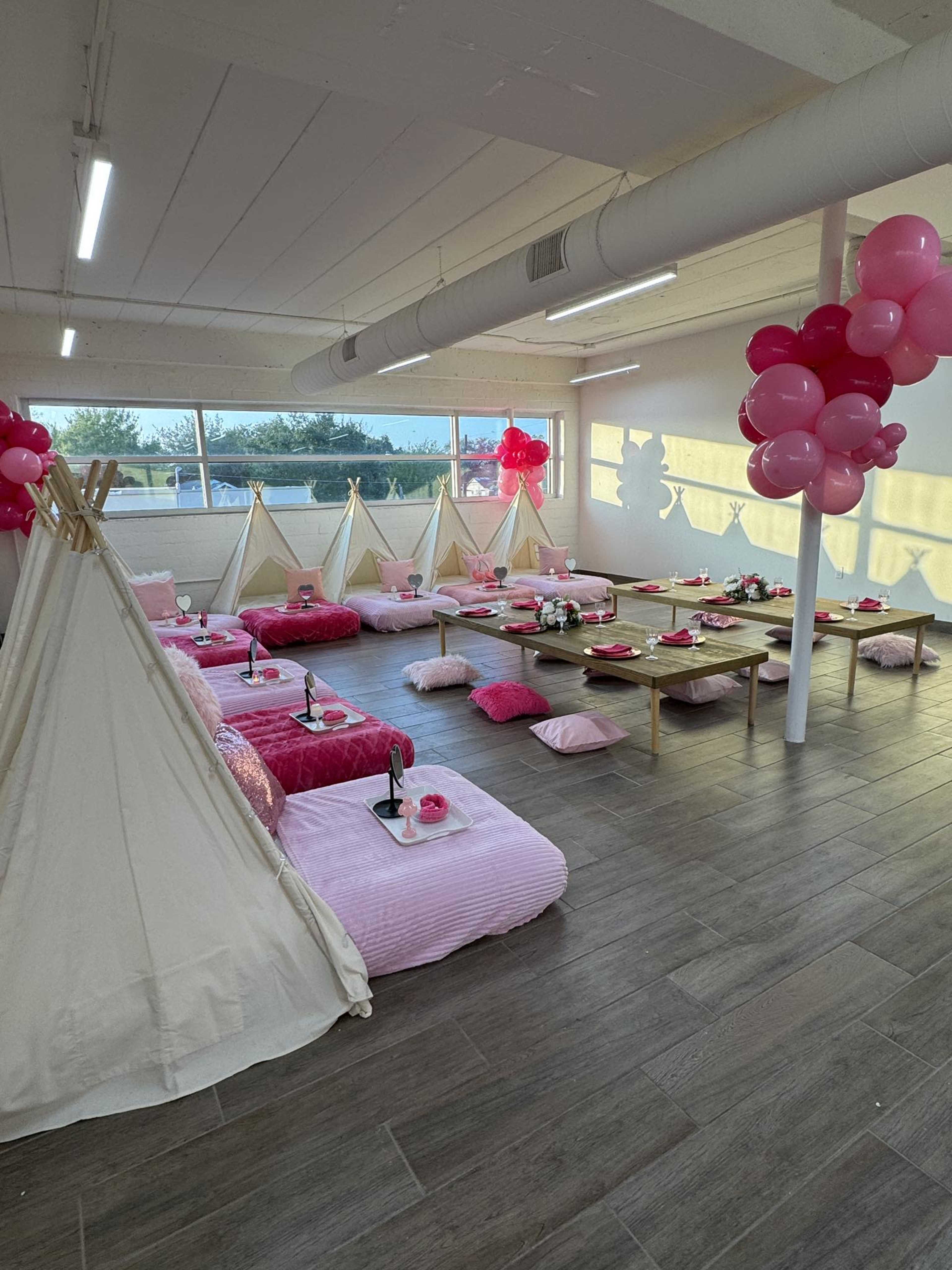 The image shows a brightly lit room set up for a party, featuring multiple teepees, pink cushions, tables with snacks, and balloon decorations.