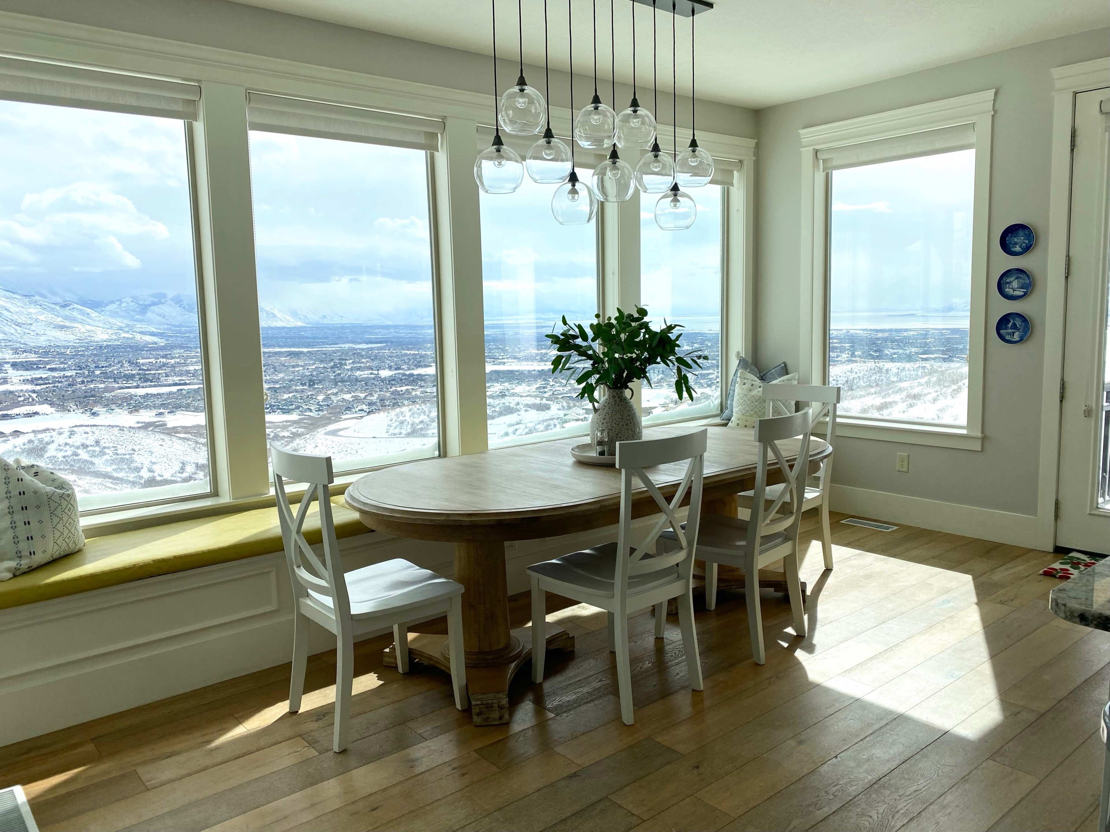 A dining area with a round wooden table surrounded by white chairs, large windows showcasing a mountain view, and a pendant light fixture above.