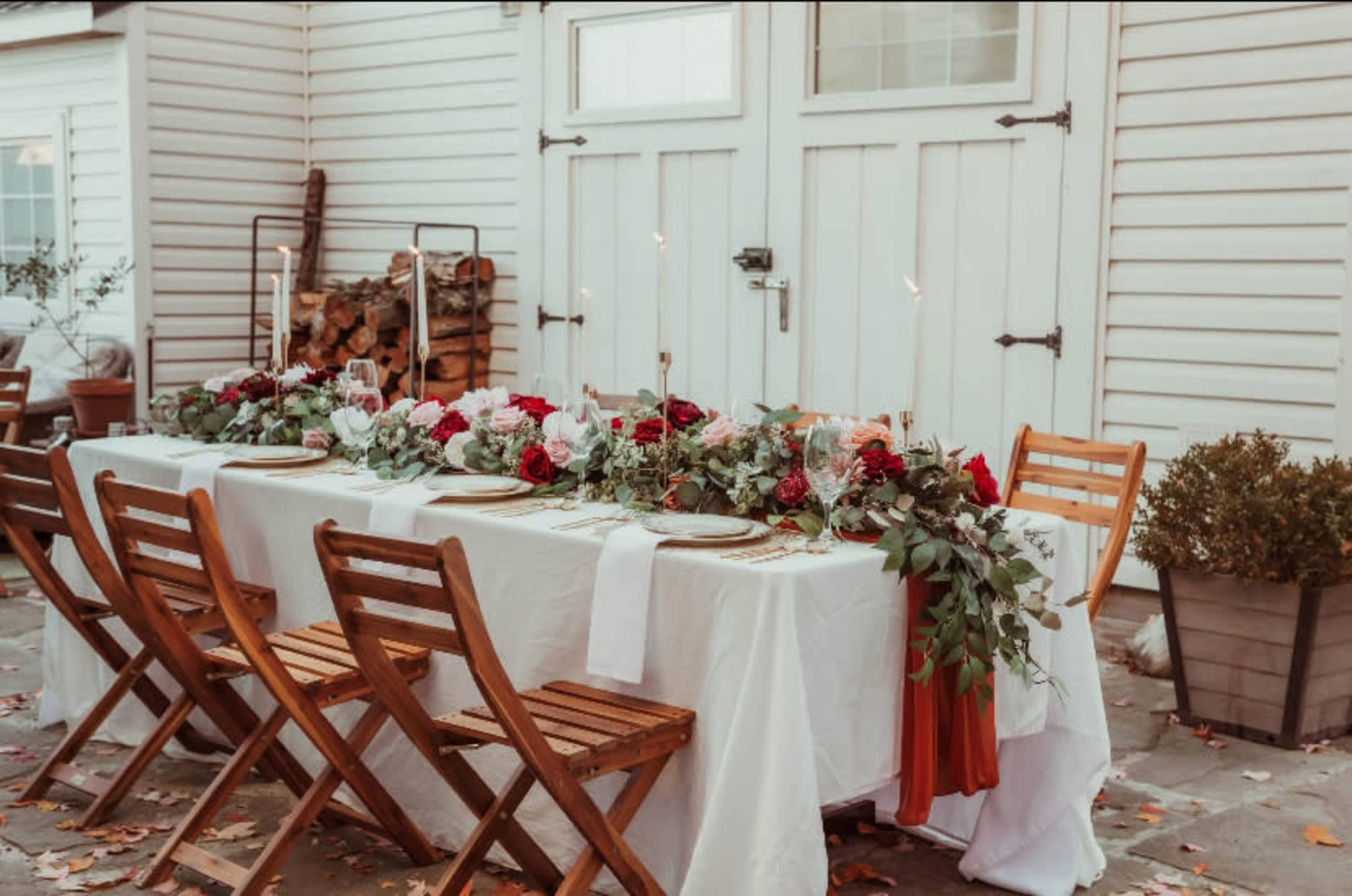 A long table is set outdoors with white linens, gold tableware, and floral decorations, situated against a backdrop of a white wooden wall and stacked firewood.