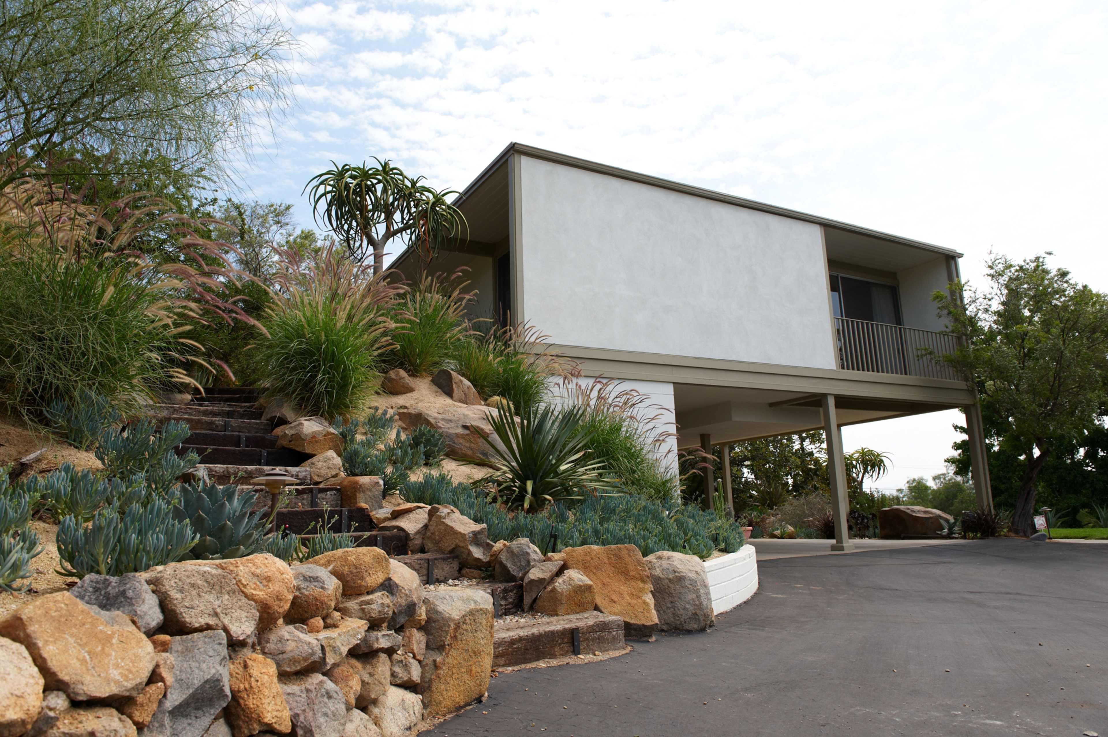 A modern two-story house is set on a landscaped hill with stone steps and various plants leading up to its entrance.