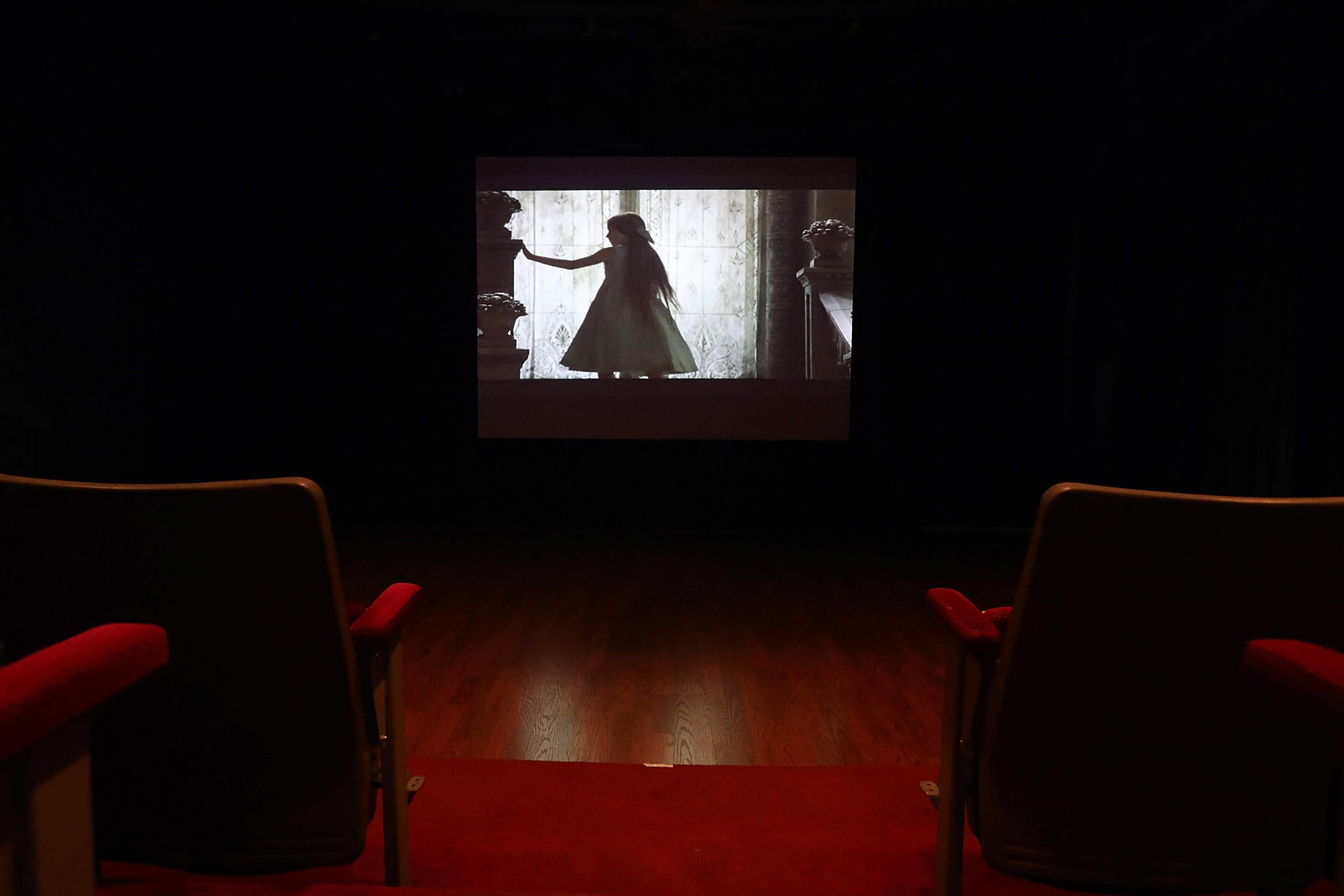 A projection screen displays a silhouette of a girl in a dress, viewed from empty red theater seats.
