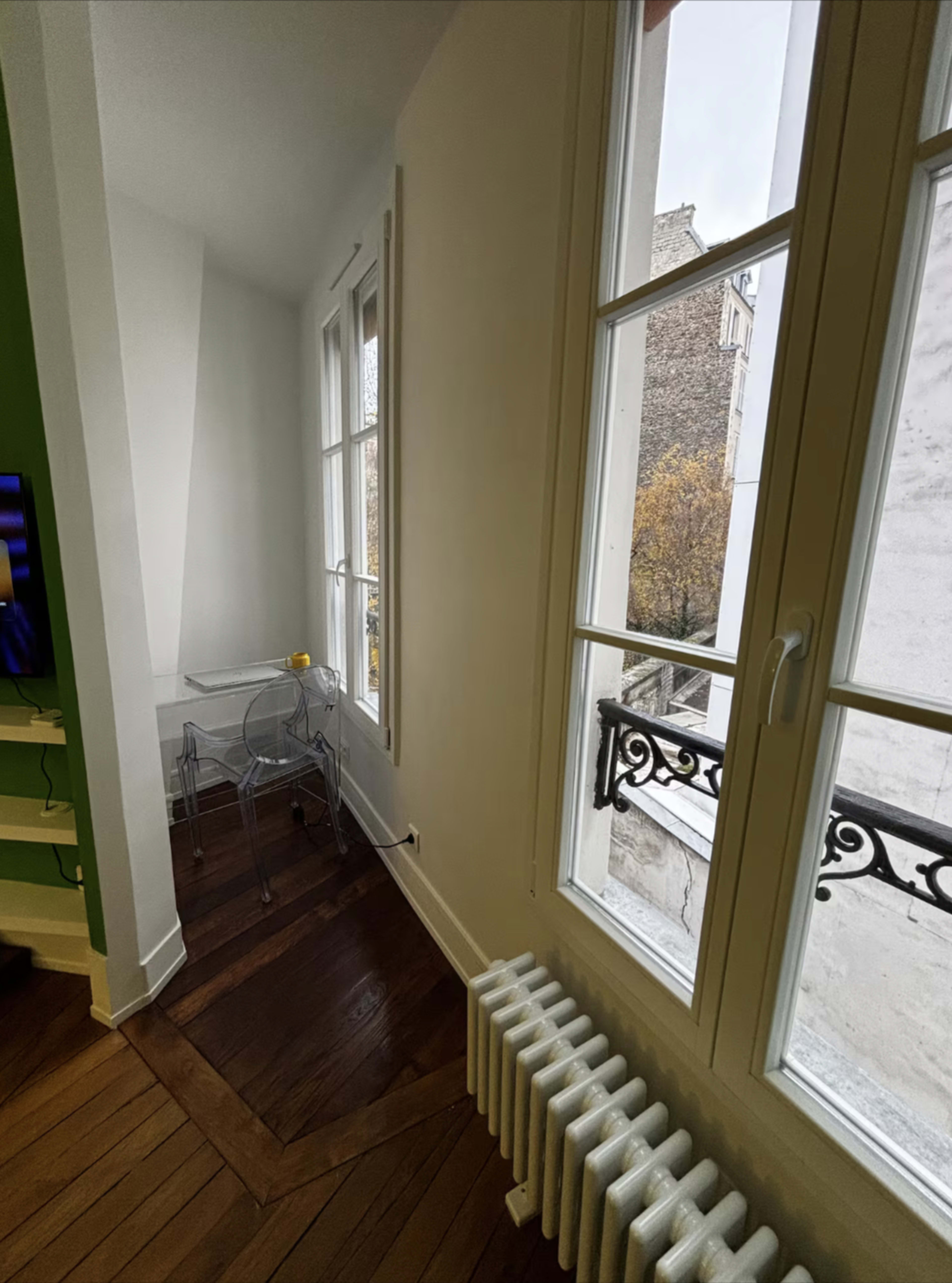 The image shows a corner of a room with large windows, a white radiator, and a transparent chair positioned near the window.