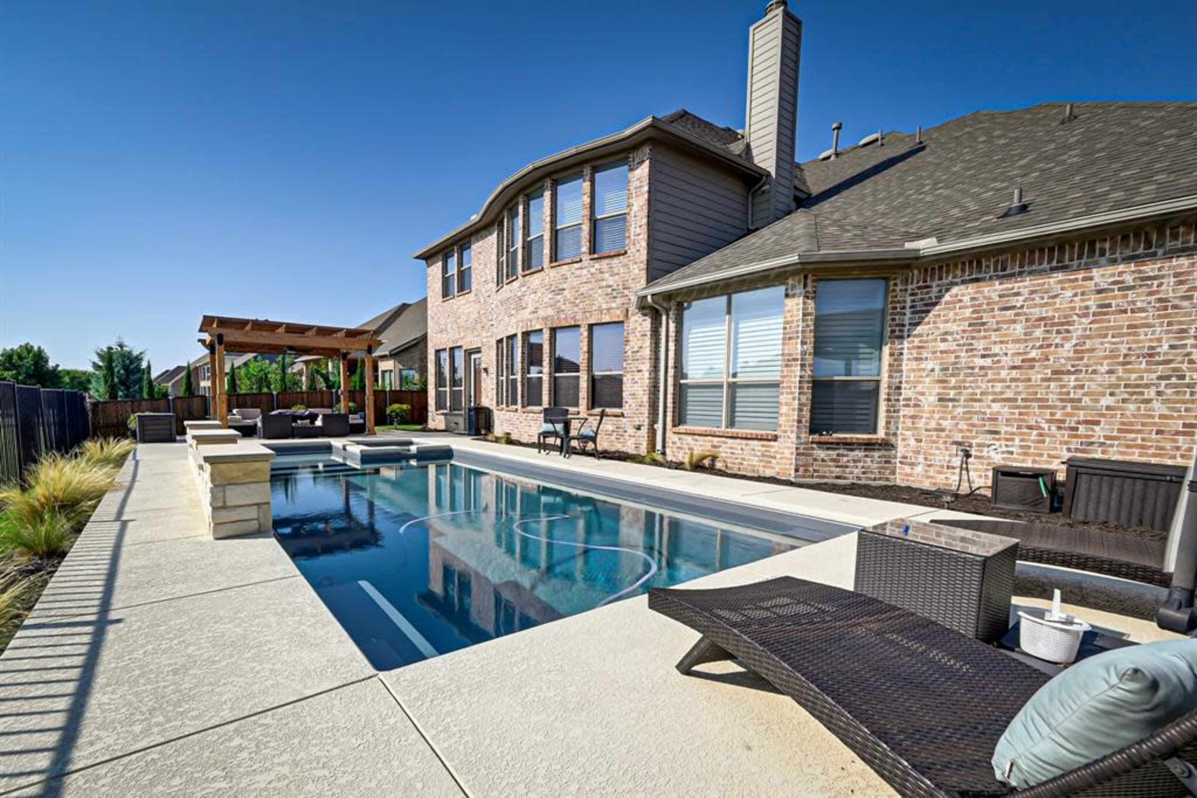 A backyard with a rectangular pool adjacent to a brick house, featuring a shaded seating area and landscaped greenery.