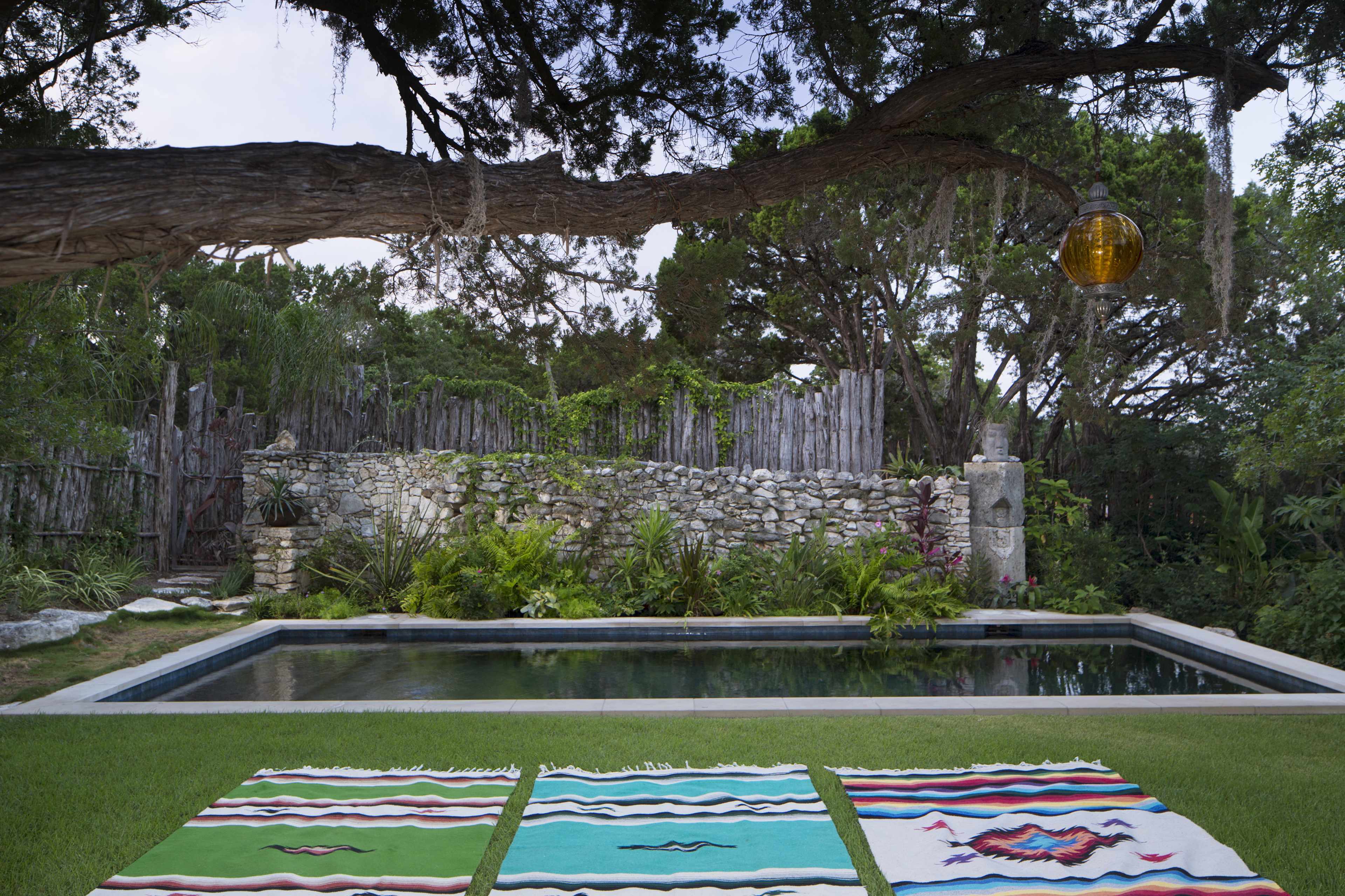A serene outdoor scene with a rectangular swimming pool surrounded by lush greenery and a stone wall, with colorful woven blankets laid out on the grass in the foreground.