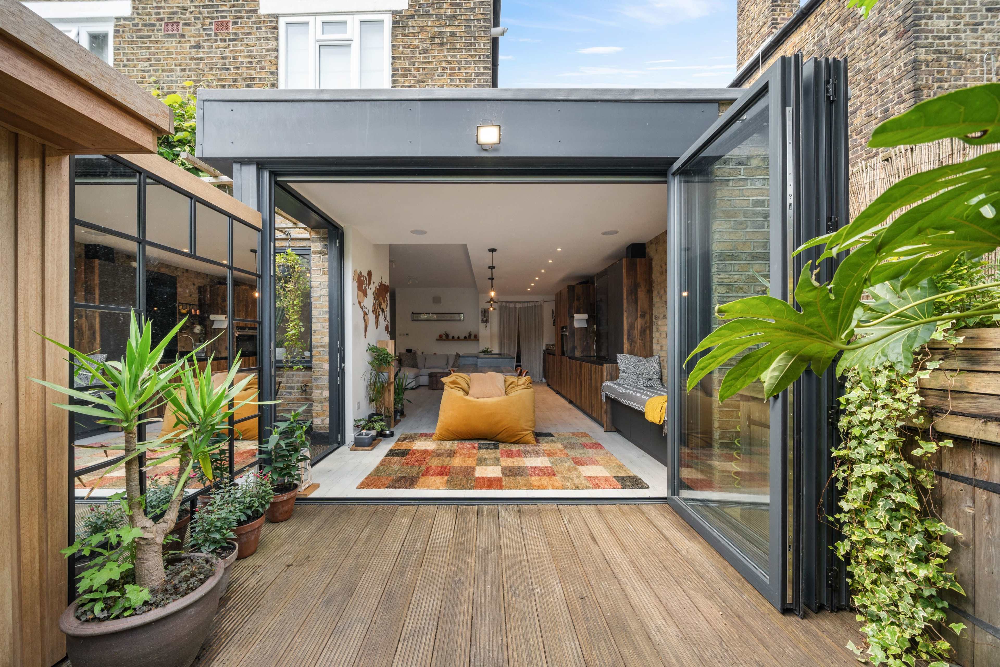 A modern living space with large glass doors open to a wooden deck surrounded by potted plants and a colorful rug inside.