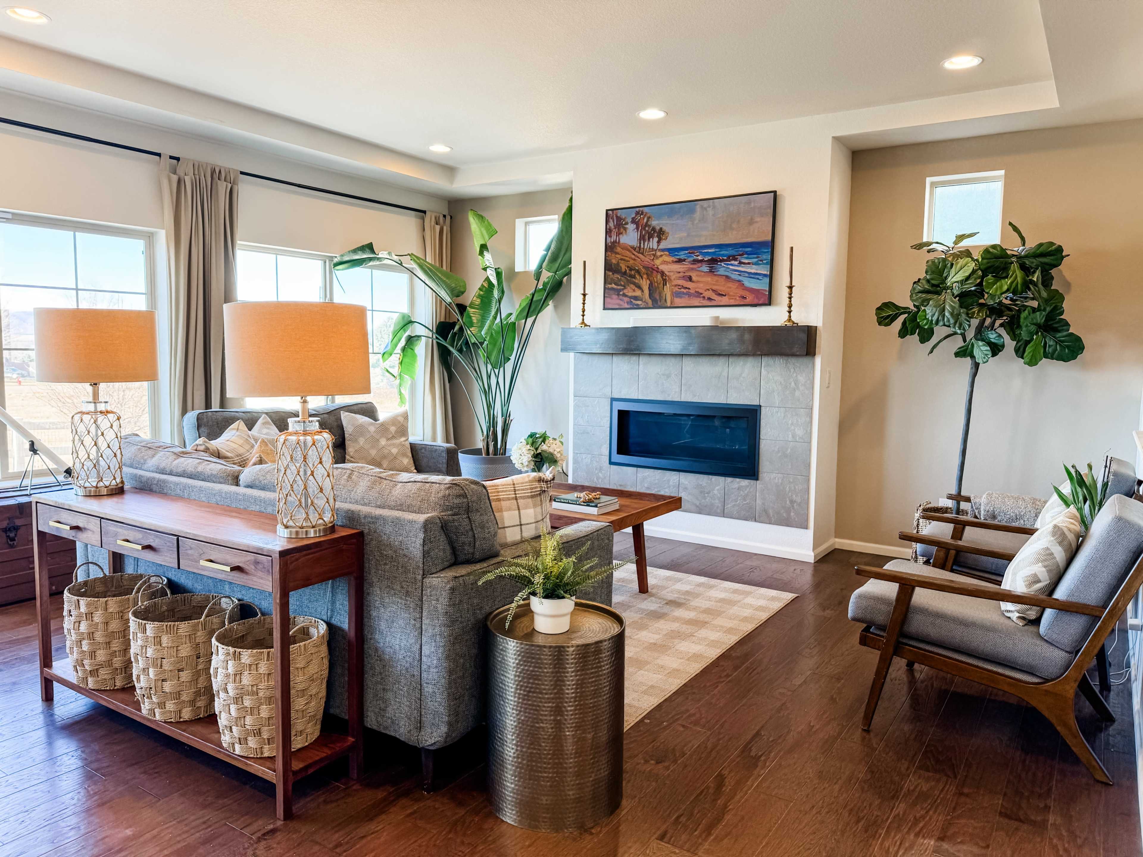 A modern living room features a gray sofa, wooden furniture, a stone fireplace, and large windows with plants and decorative items.
