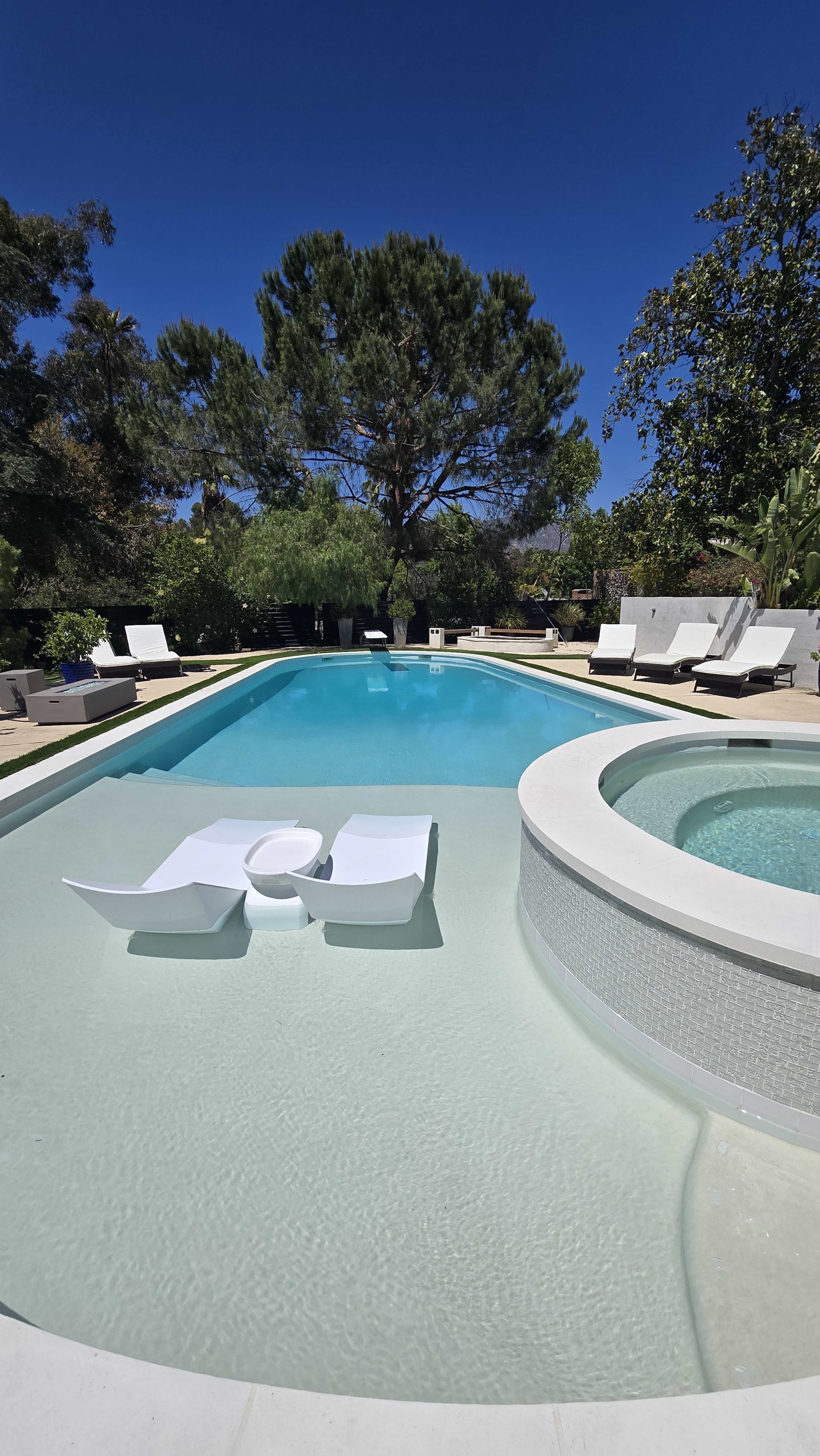 A modern swimming pool with a circular hot tub and white lounge chairs surrounds it, set against a clear blue sky and greenery.