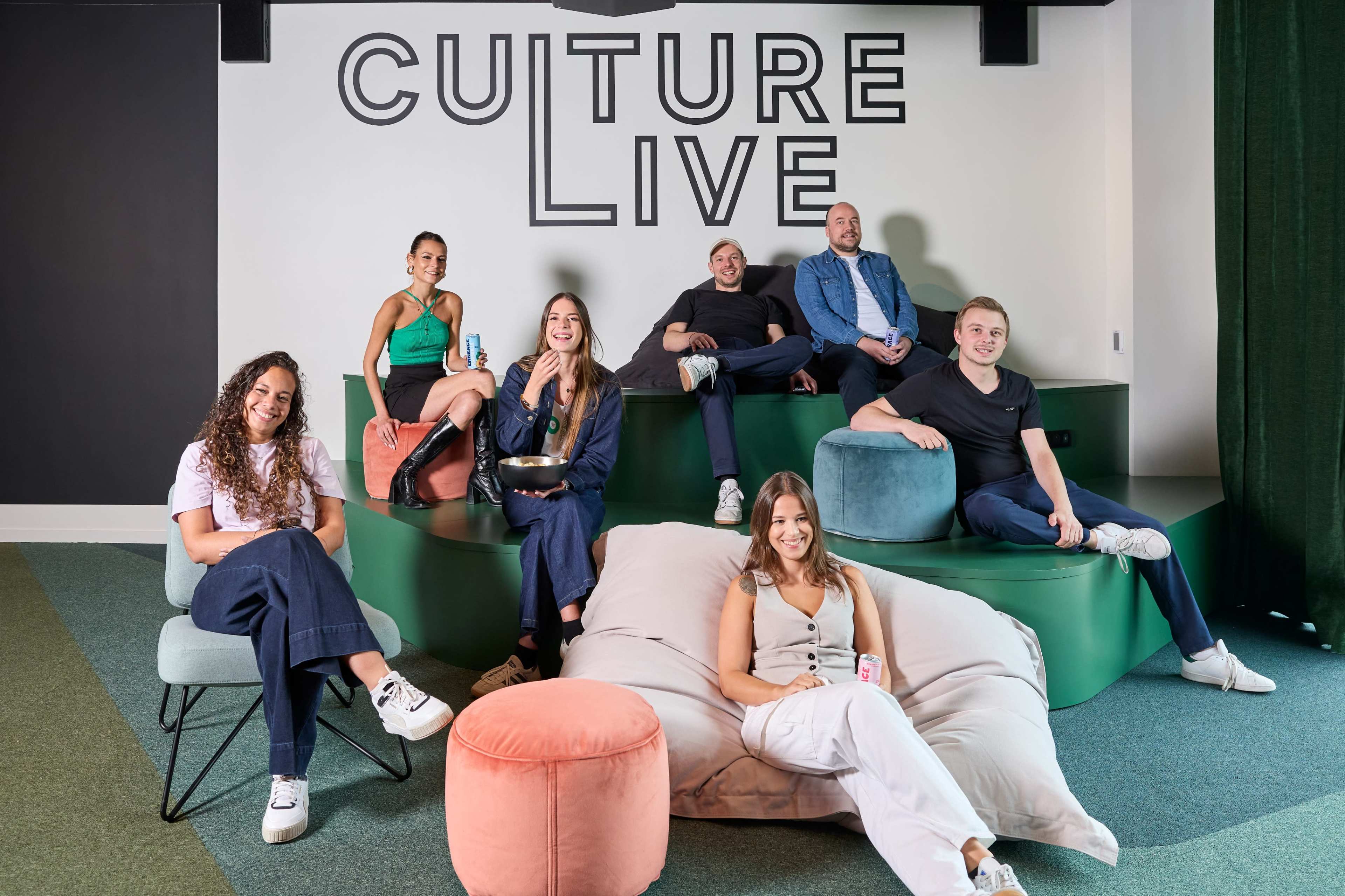 A group of seven people is seated on colorful furniture in a modern space with a large "CULTURE LIVE" sign on the wall behind them.