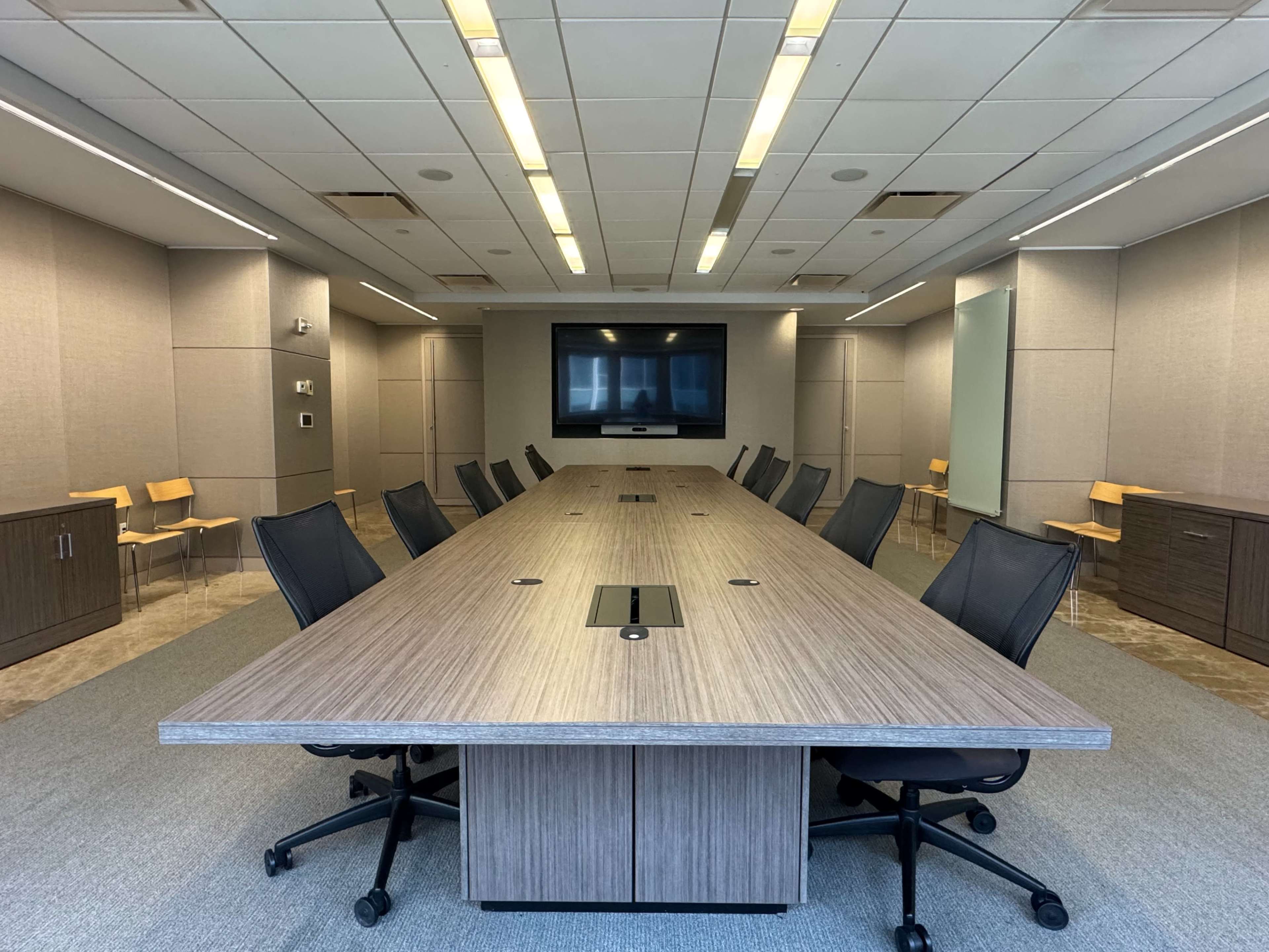 The image shows a modern conference room with a long rectangular table, black office chairs, and a large screen mounted on the wall.
