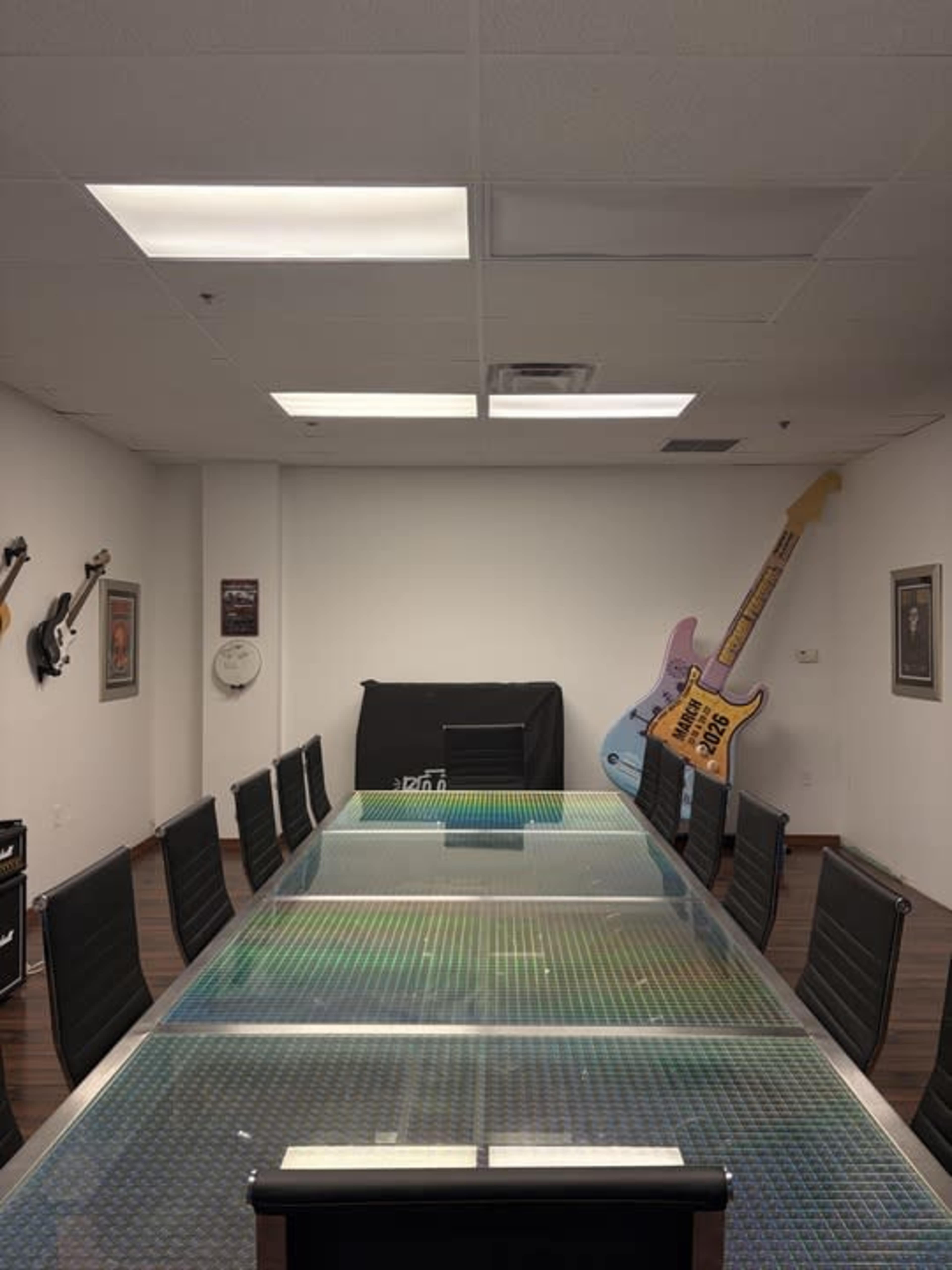 A long glass conference table is set in a room with black chairs, two electric guitars mounted on the walls, and a piano in the background.