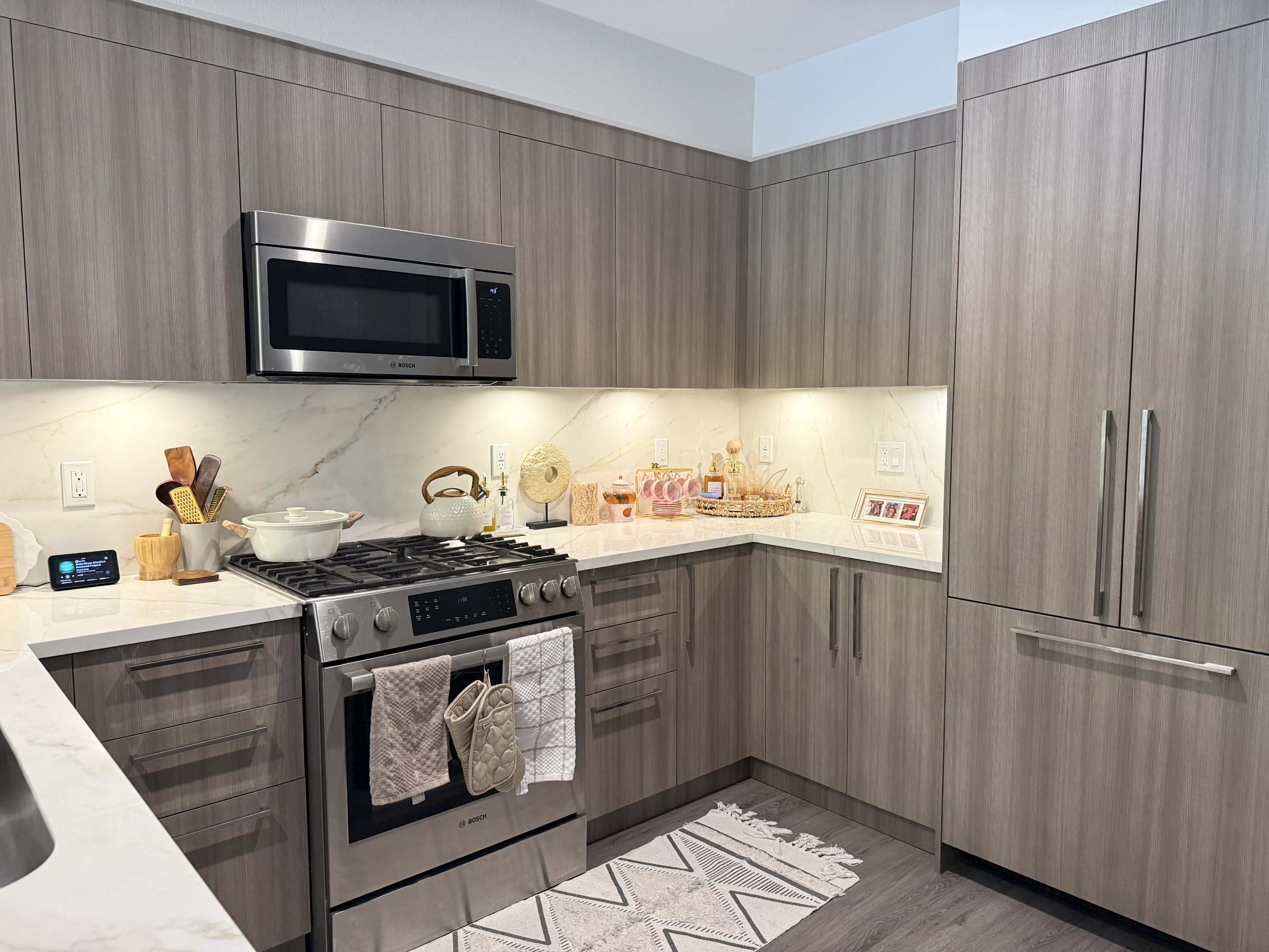The image shows a modern kitchen featuring gray cabinetry, a stainless steel microwave, and a gas stove with a white marble countertop and a patterned rug.