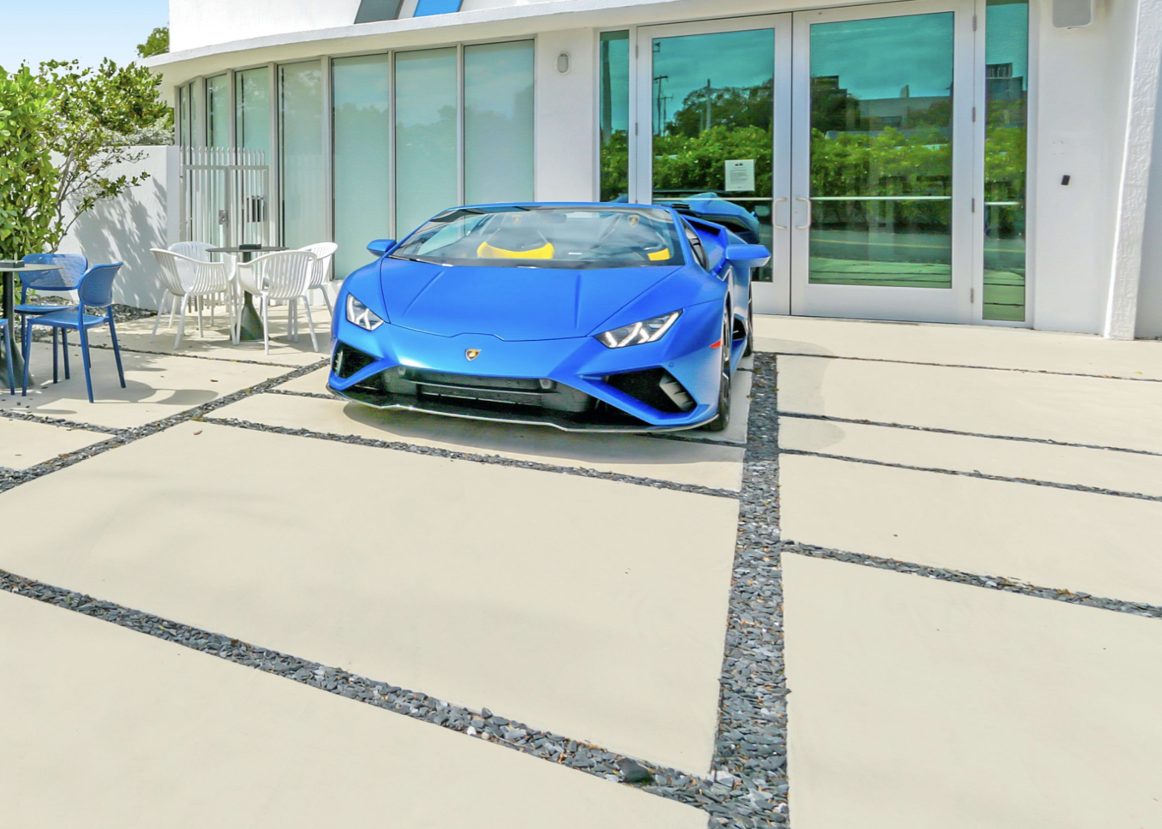 A blue Lamborghini is parked on a concrete pathway in front of a modern building with large glass doors.
