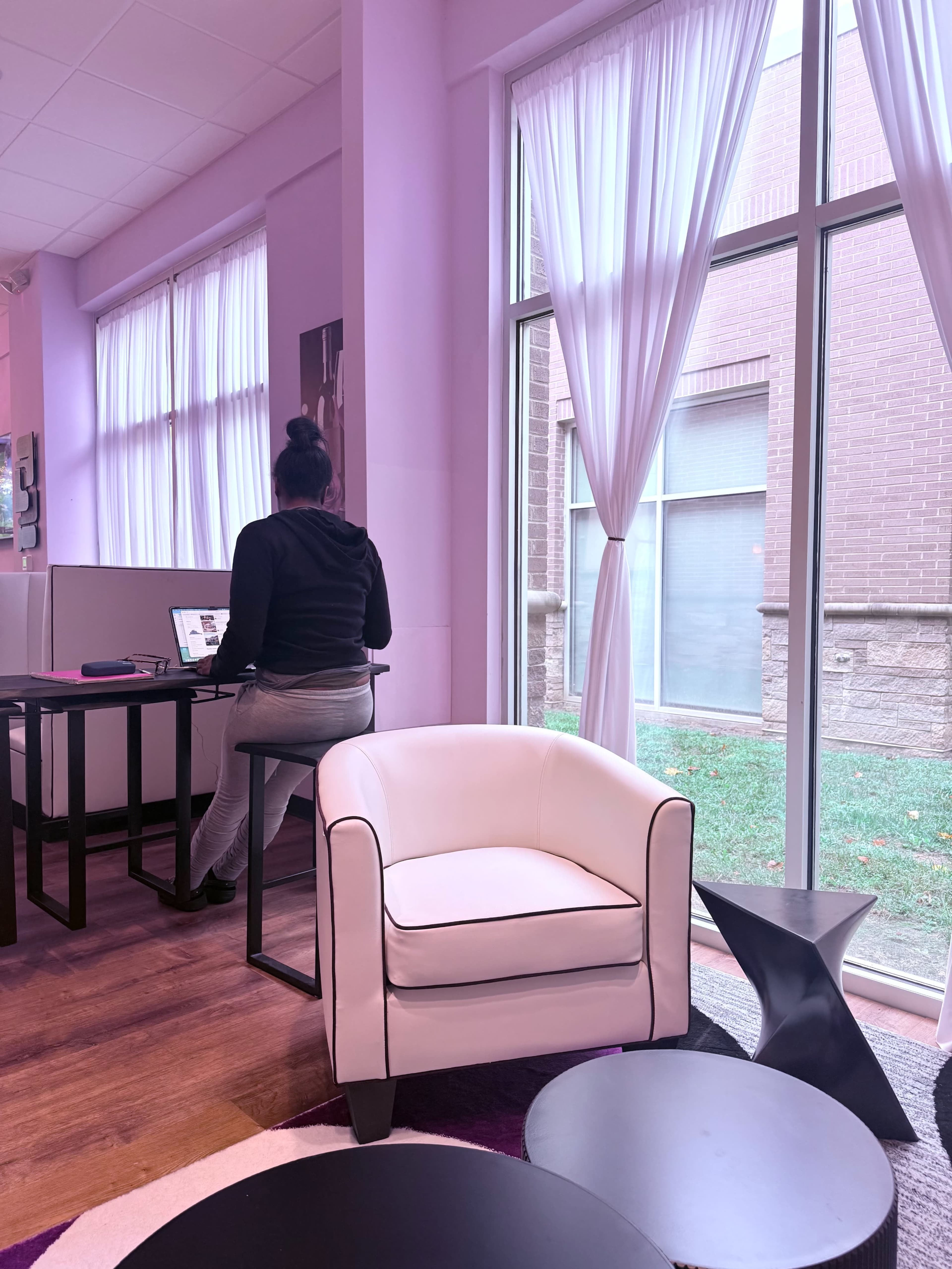 A person sits at a desk with a computer near large windows adorned with white curtains, while a white armchair and small black tables are positioned in the foreground.