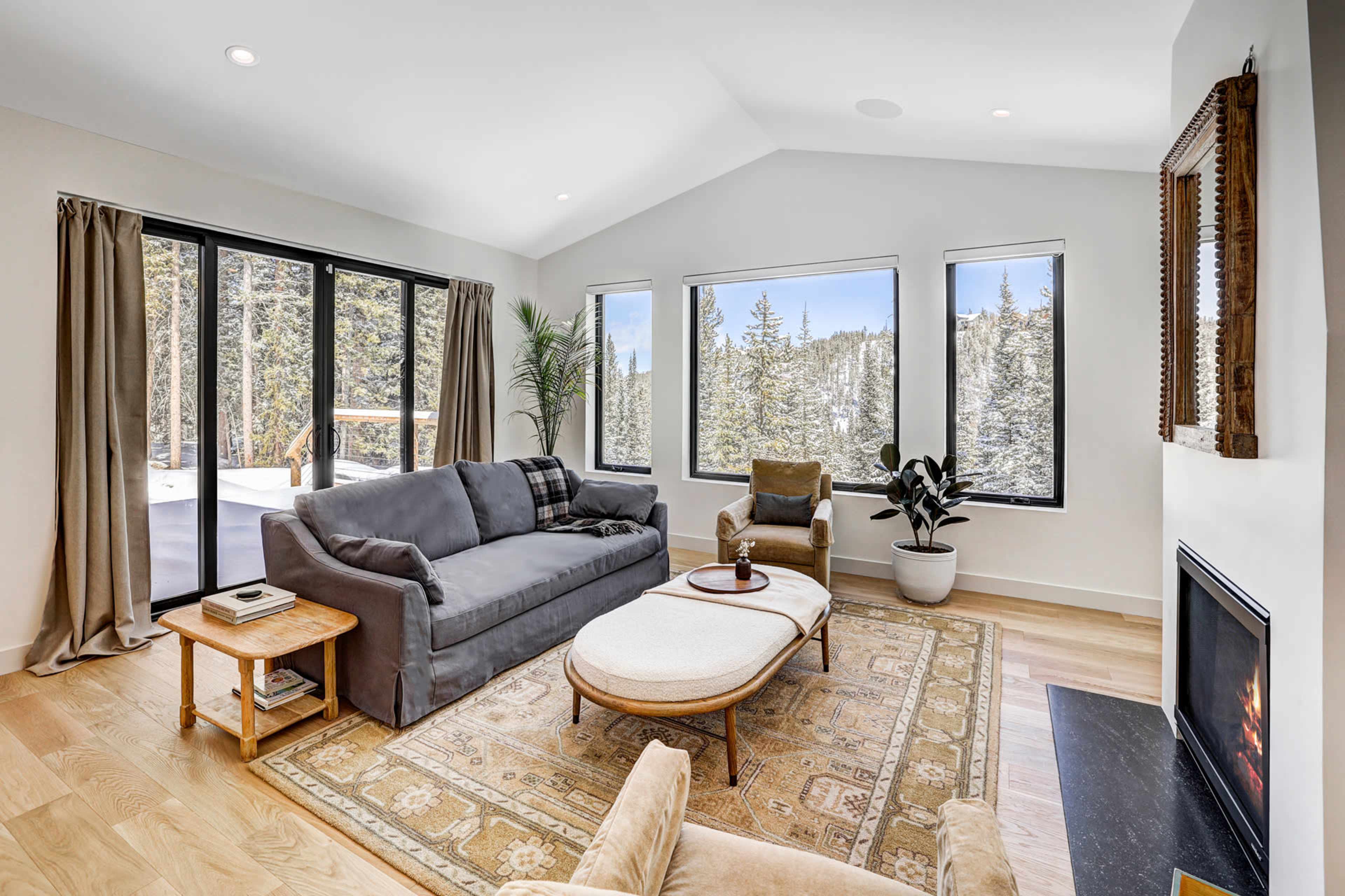 A modern living room features large windows overlooking a snowy landscape, with a sofa, armchair, and a coffee table on a patterned rug.