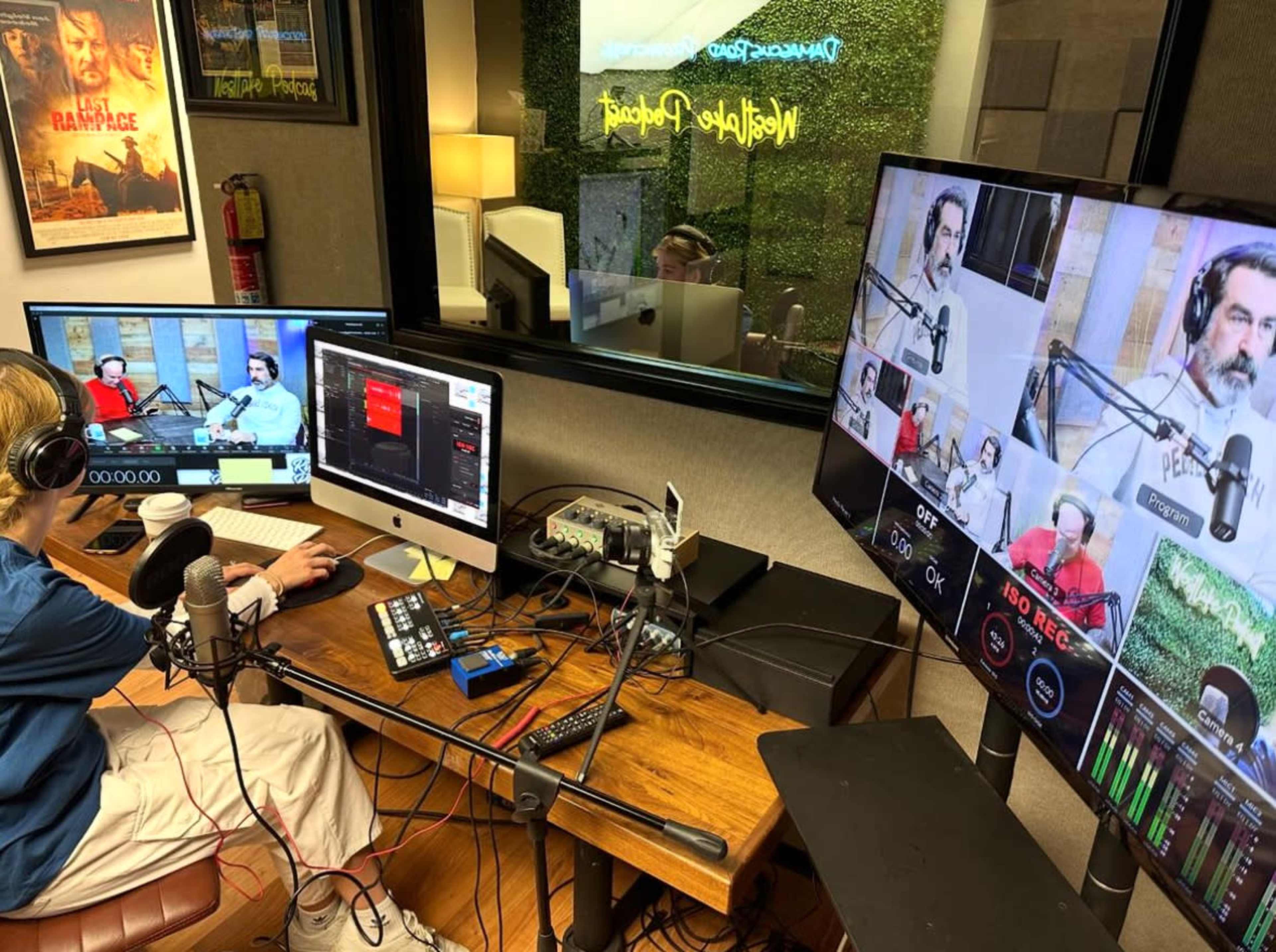 A person sits at a desk with multiple monitors and audio equipment, while engaging in a recording session in a well-lit studio.