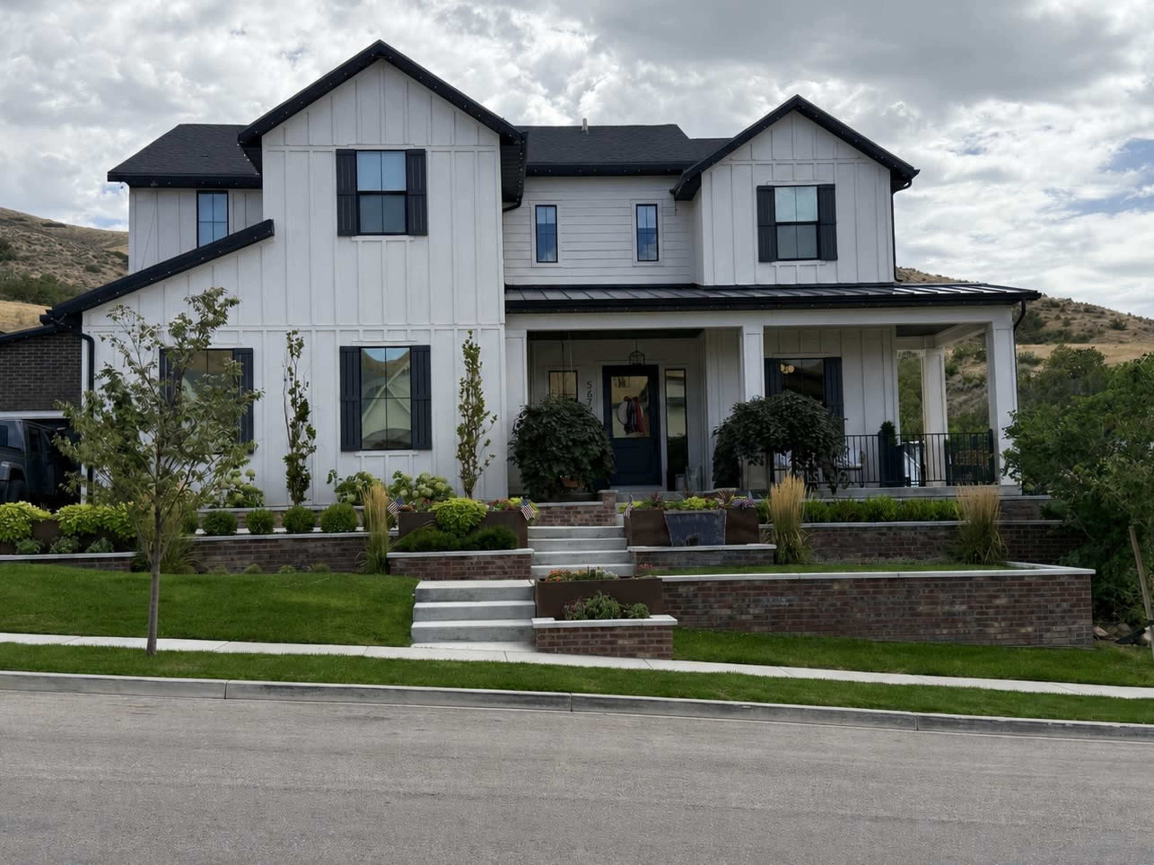 The image shows a two-story house with a mix of white and dark exterior features, surrounded by landscaped greenery and stone pathways.