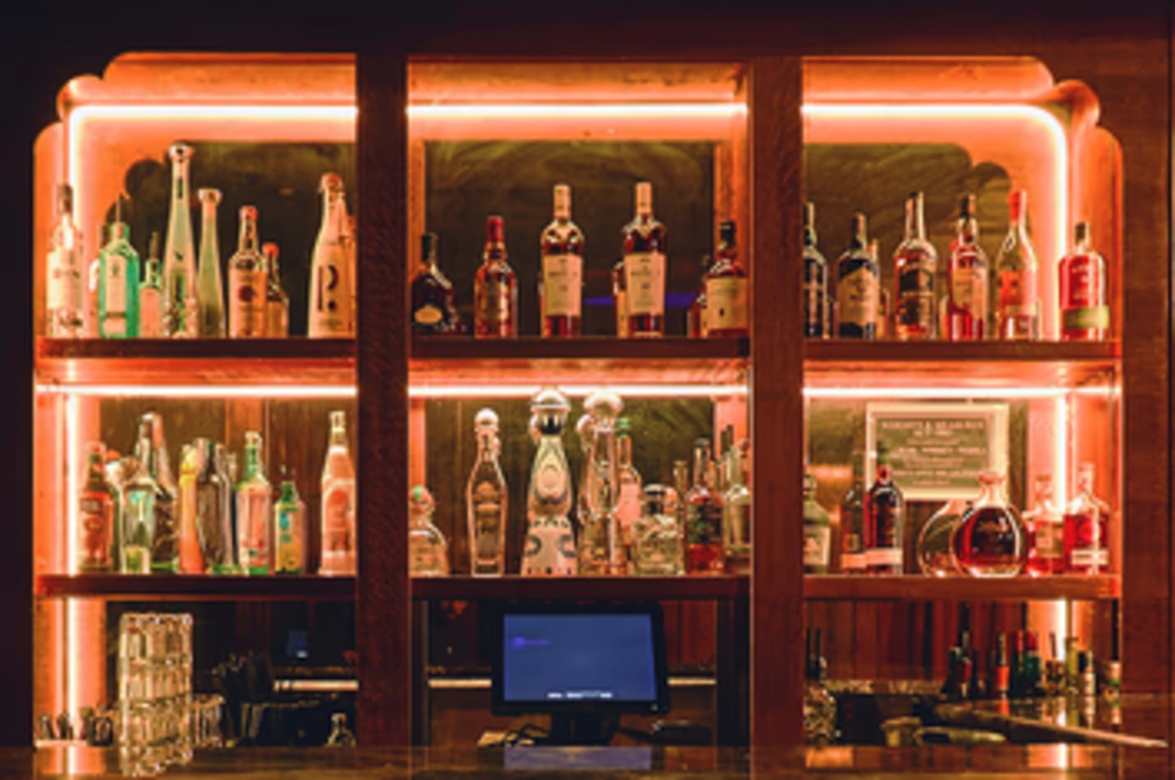 The image shows a well-organized bar counter with numerous bottles of liquor displayed on shelves, illuminated by soft backlighting.