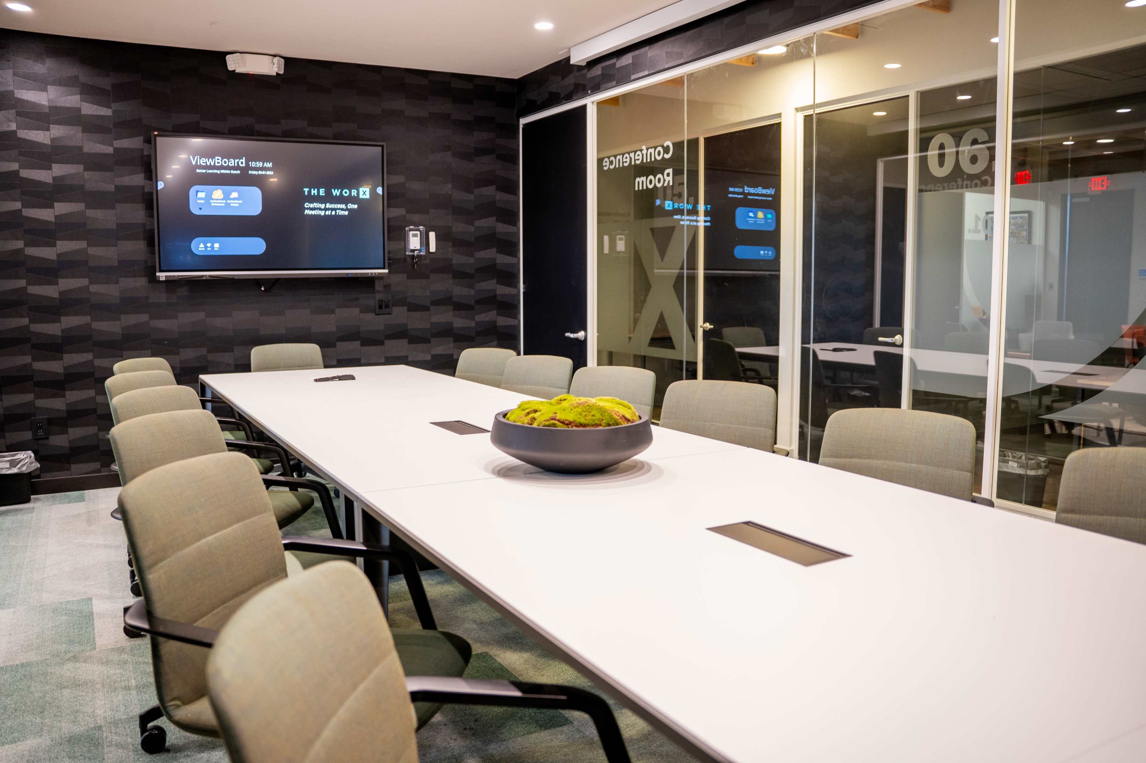 A modern conference room features a long, white table surrounded by gray chairs and a wall-mounted screen displaying a digital interface.