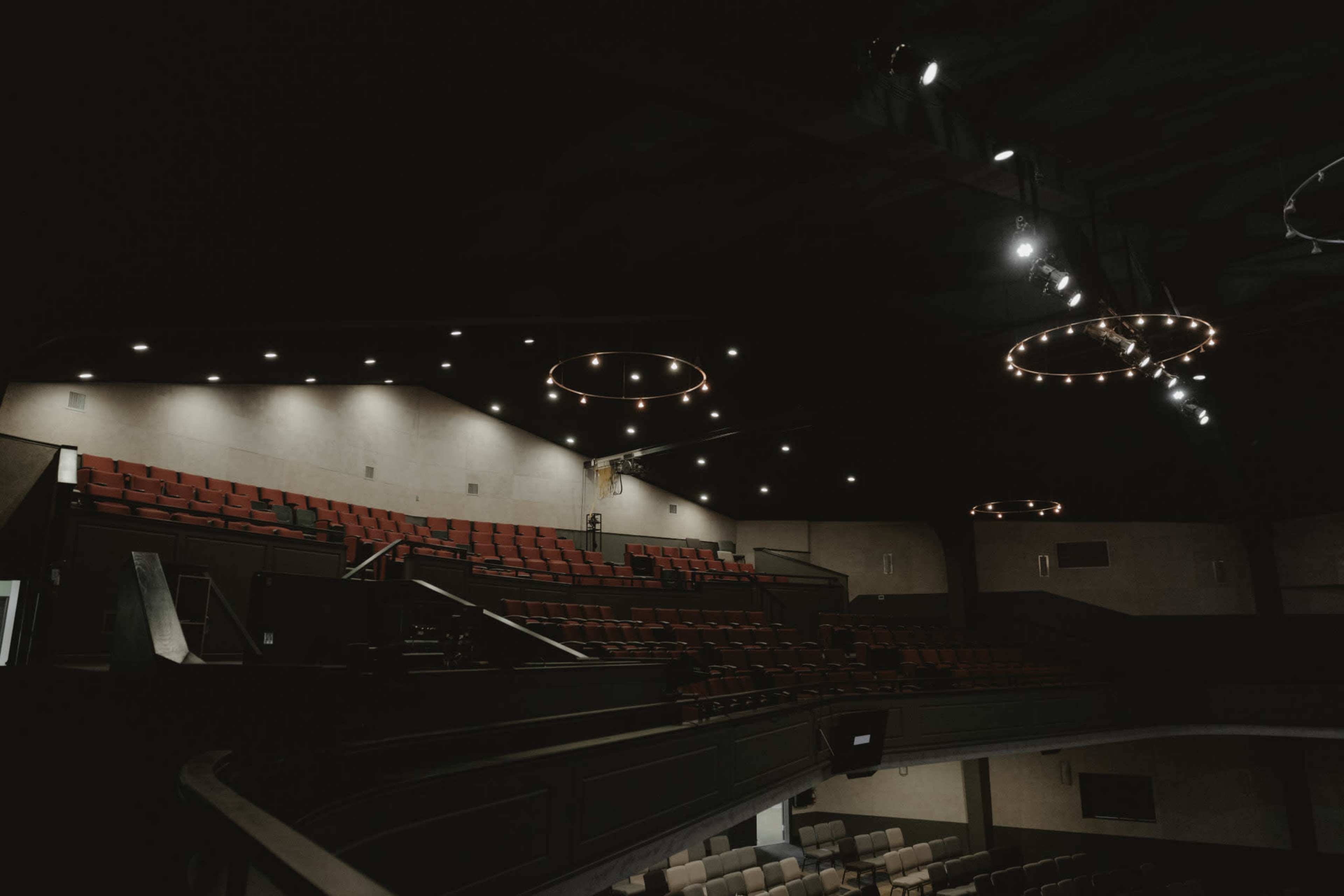 The interior of a theater features empty red seats arranged in a tiered format under circular ceiling lights.