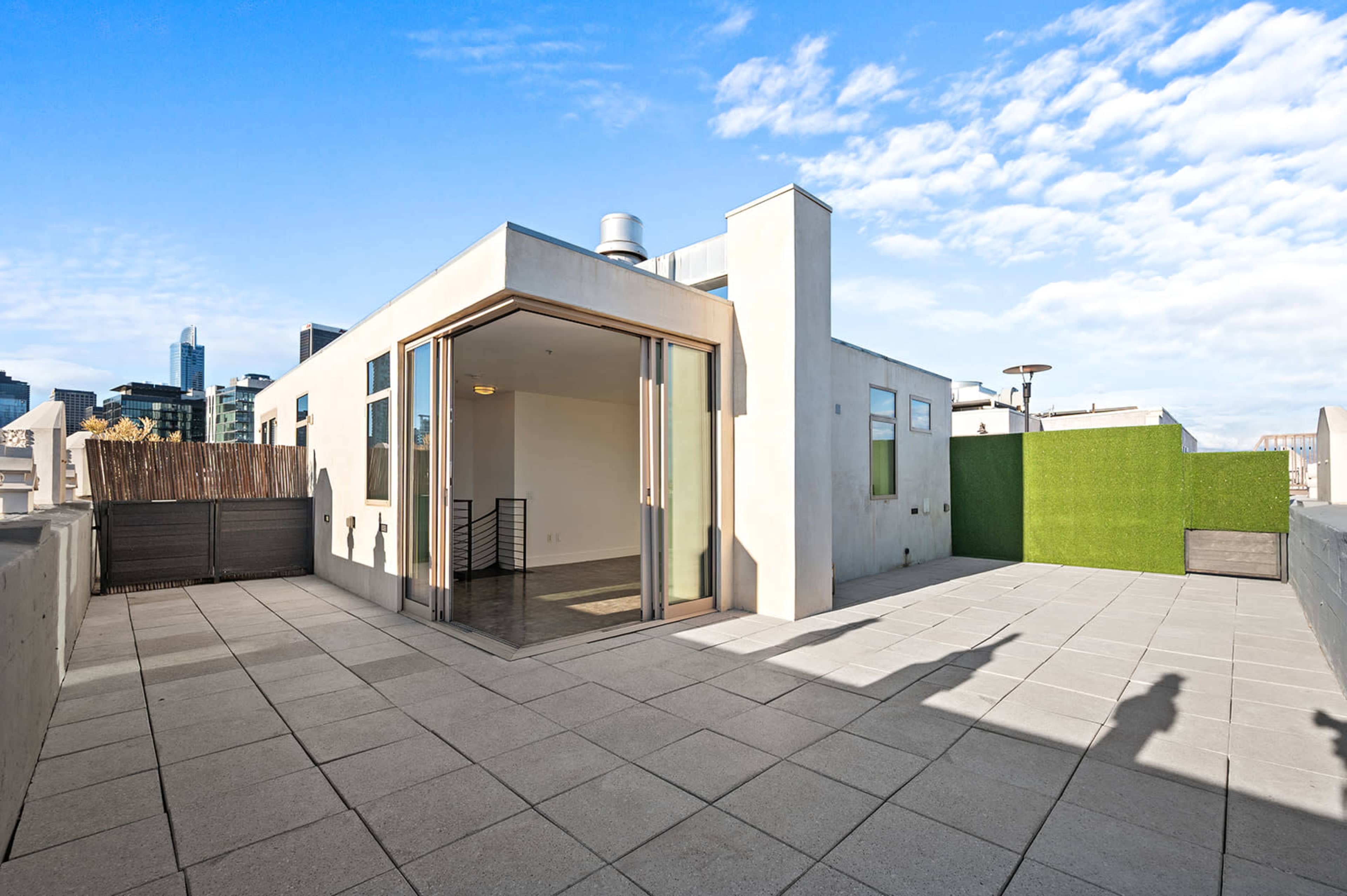 The image shows a spacious rooftop terrace featuring sliding glass doors, a green wall, and a paved surface with city buildings in the background.