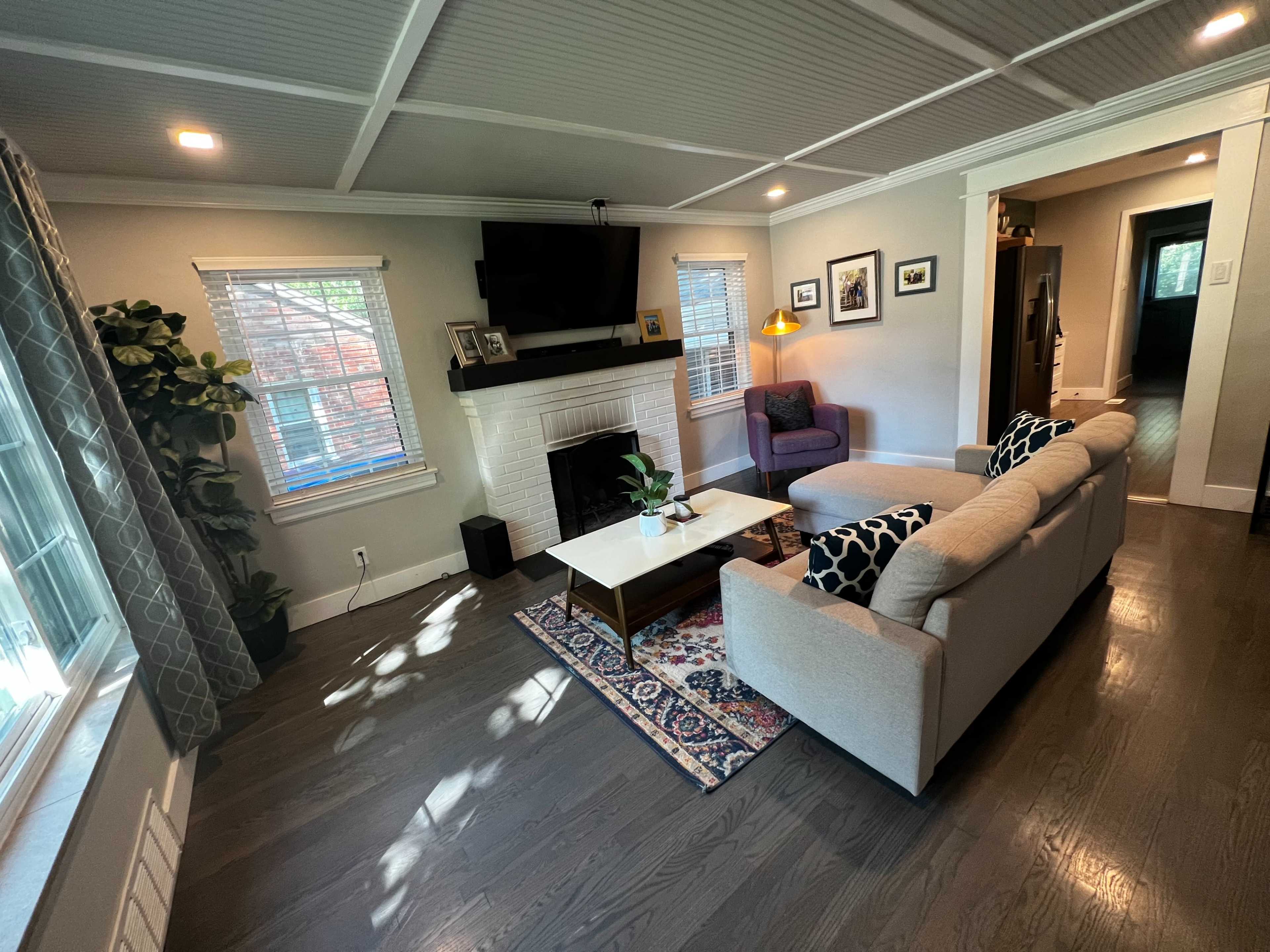 A living room features a sofa, coffee table, a wall-mounted TV above a white fireplace, and a purple armchair near a window with potted plants.