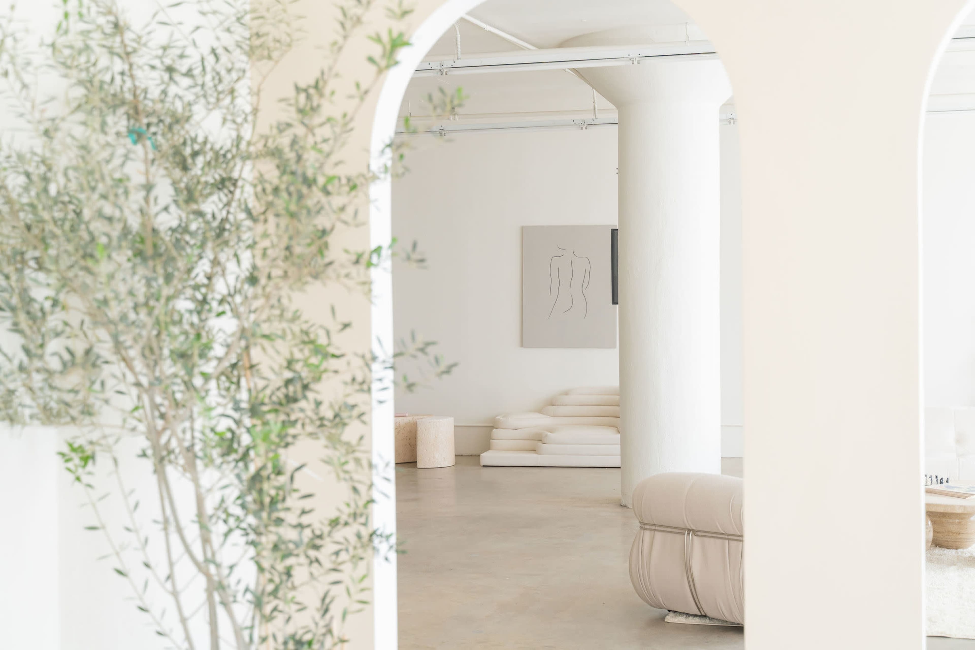 A minimalist room with a large white column, soft seating, and a piece of abstract artwork on the wall, framed by an archway and decorative greenery.
