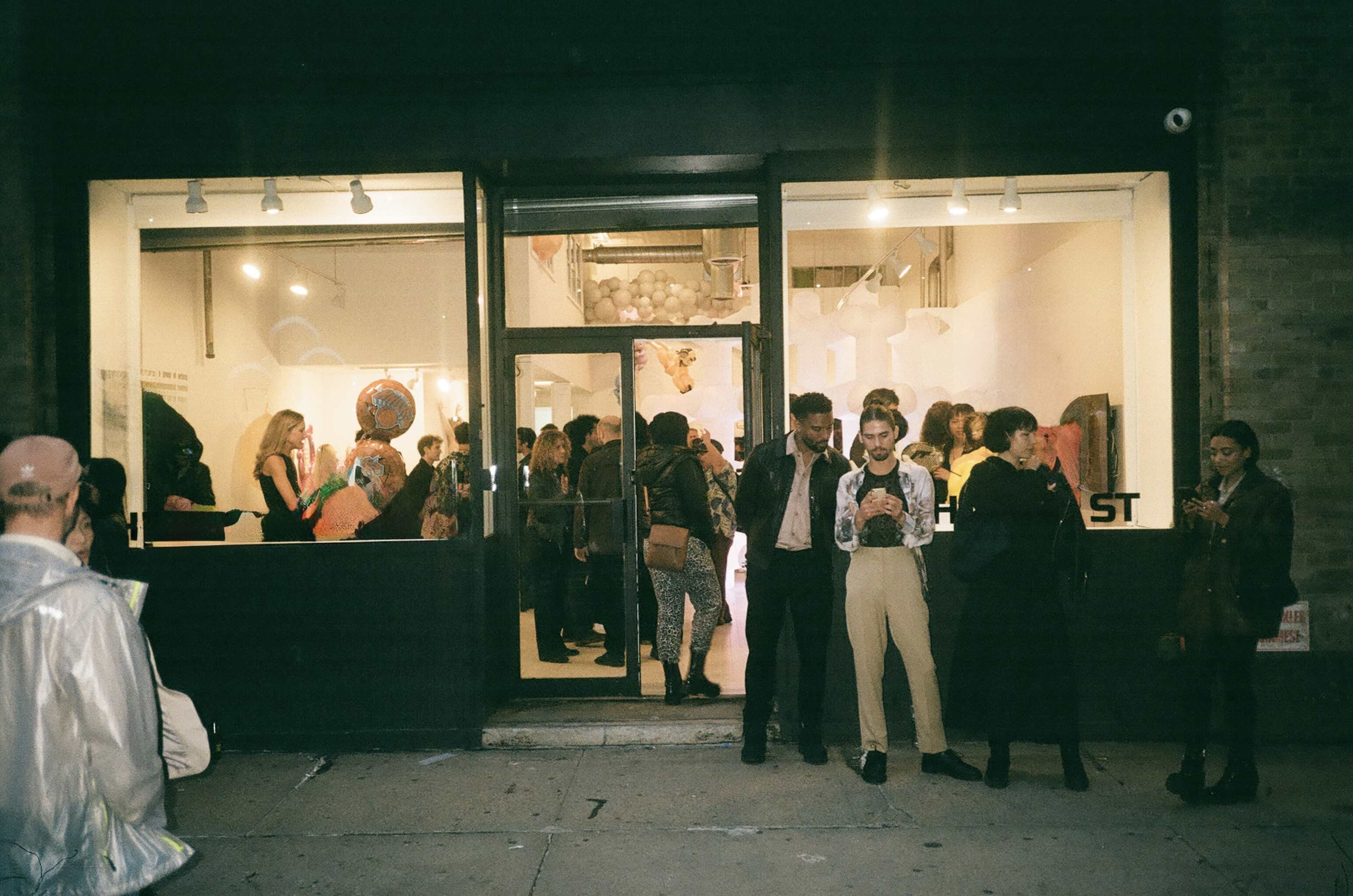 A crowd of people gathers outside an art gallery during an event, with some individuals posed in front of the entrance.
