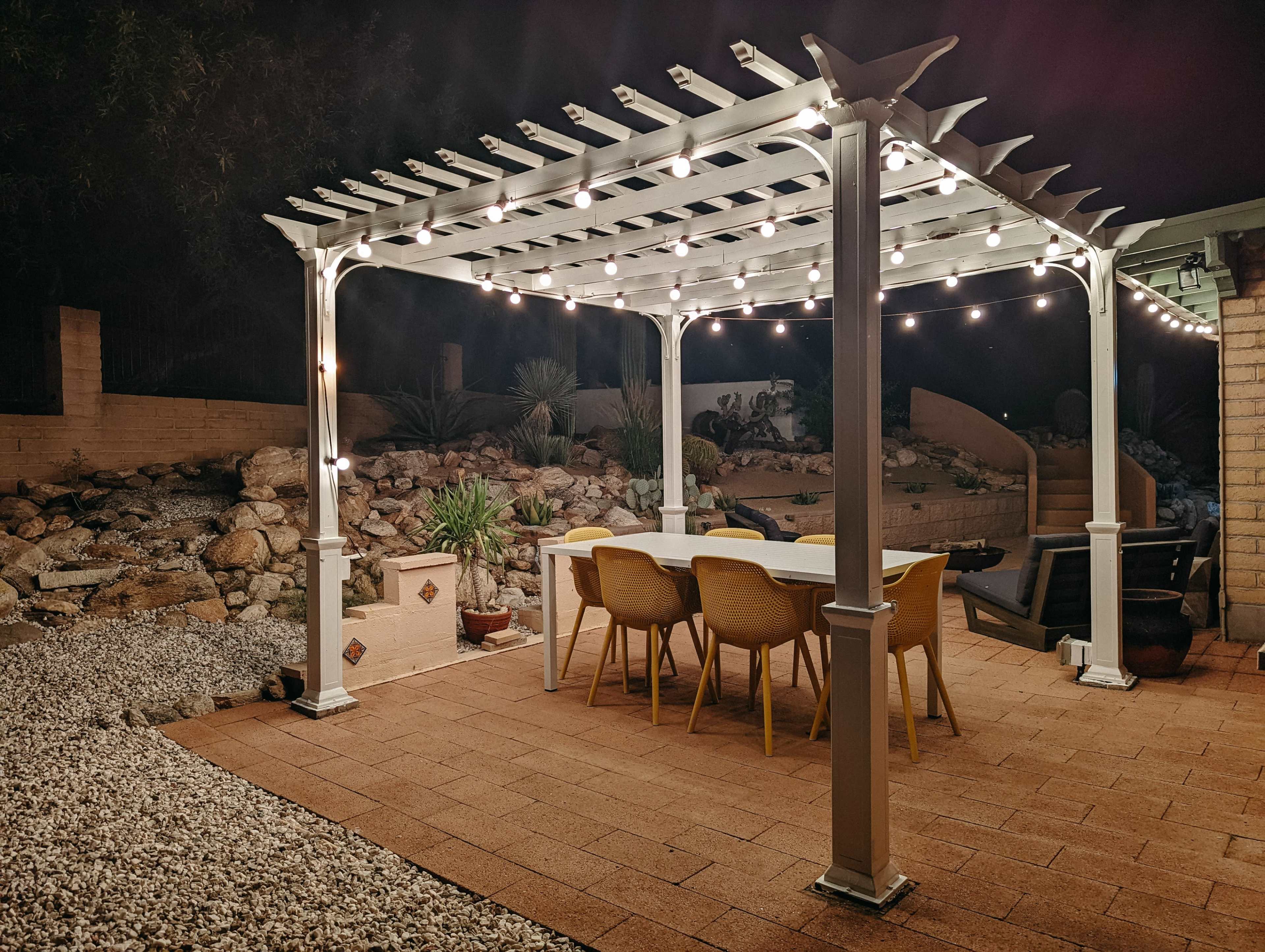 A patio area features a white pergola with string lights, surrounding a rectangular dining table and chairs, situated next to a rock garden.