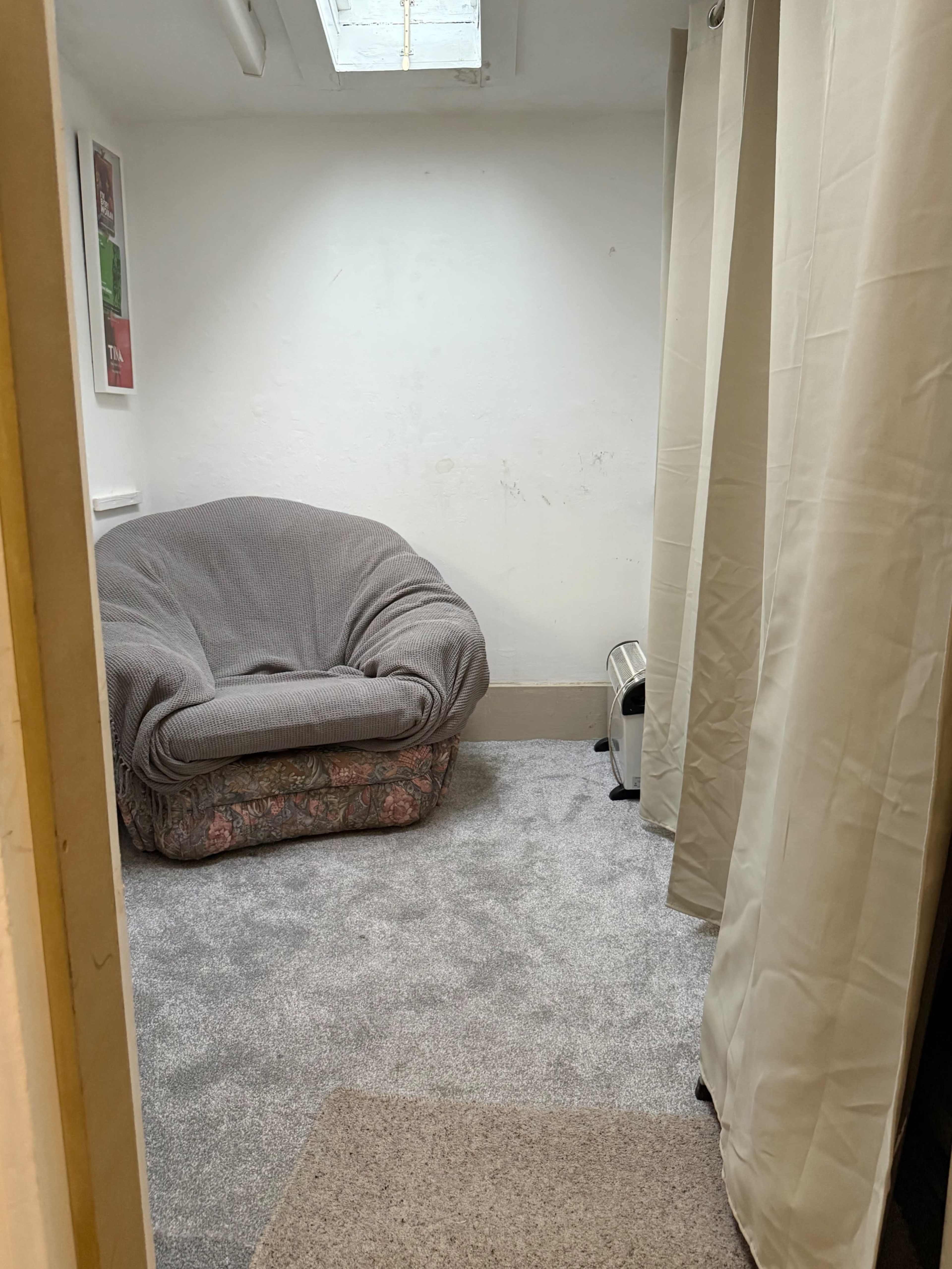 The image shows a small, sparsely furnished room with a large, comfortable chair, carpeted flooring, and light-colored curtains.