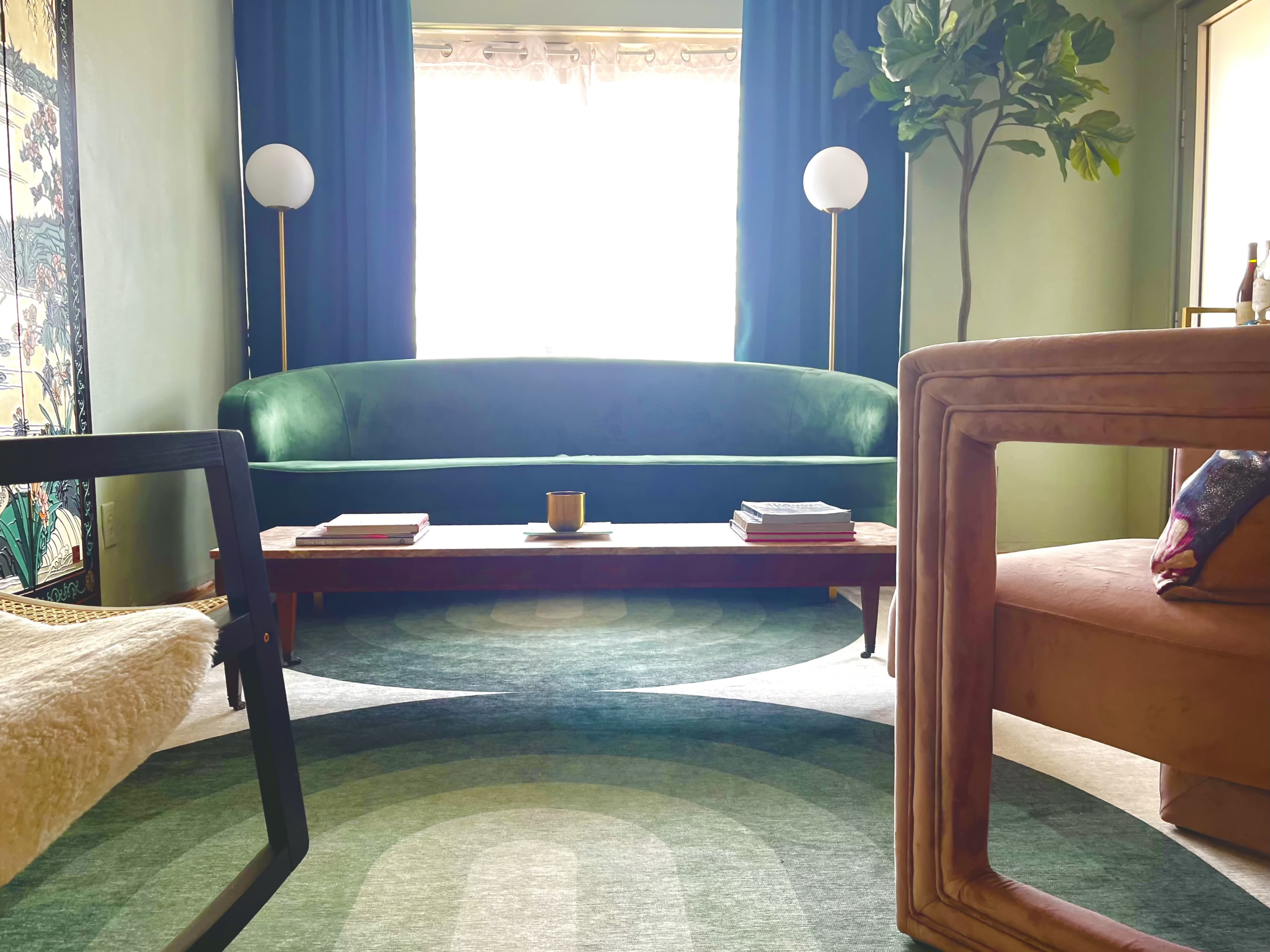 A living room with a green sofa, a coffee table with books, and large windows draped with curtains.
