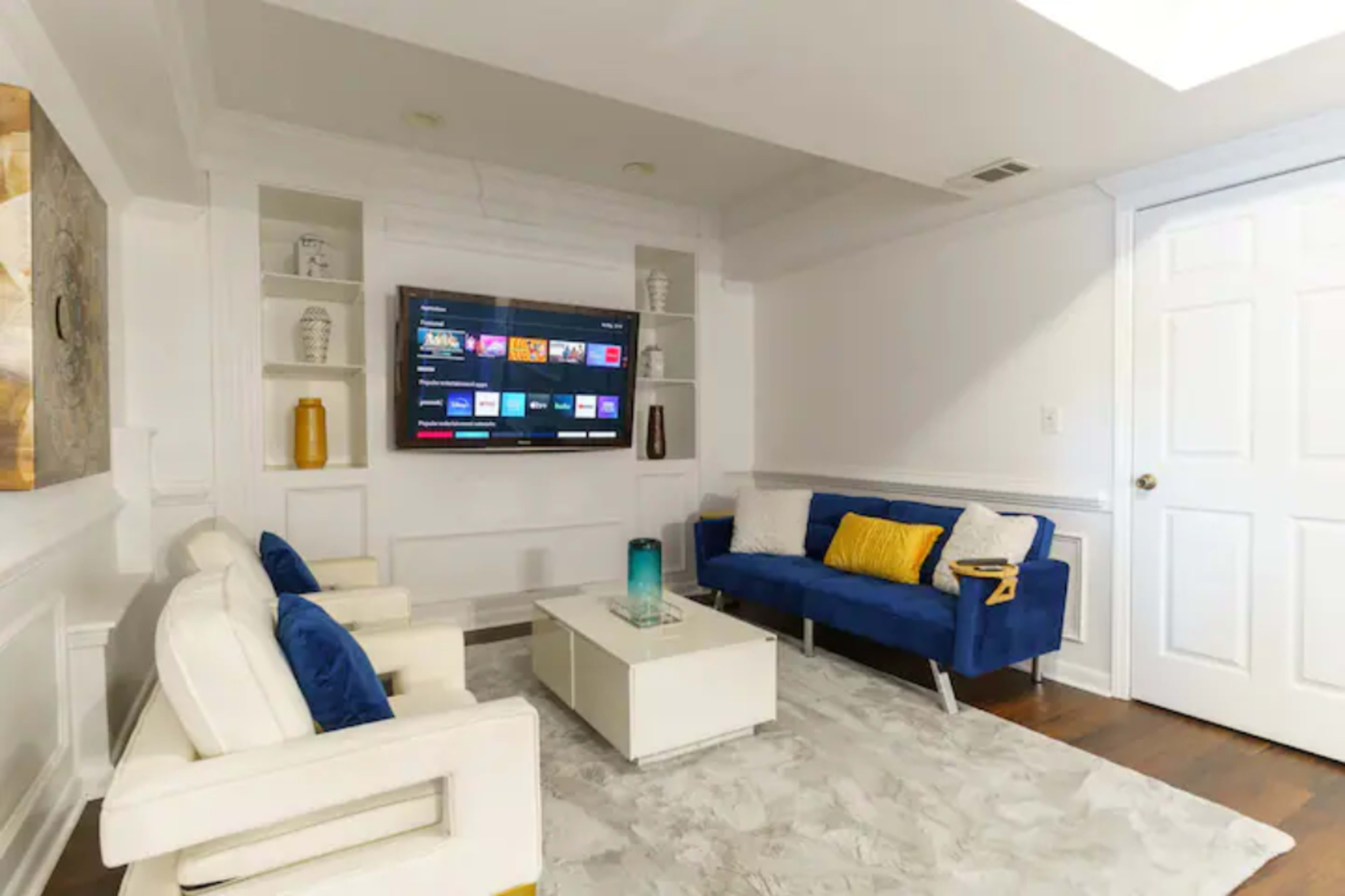 The image shows a modern living room with a blue sofa, two white chairs, a coffee table, and a wall-mounted television.