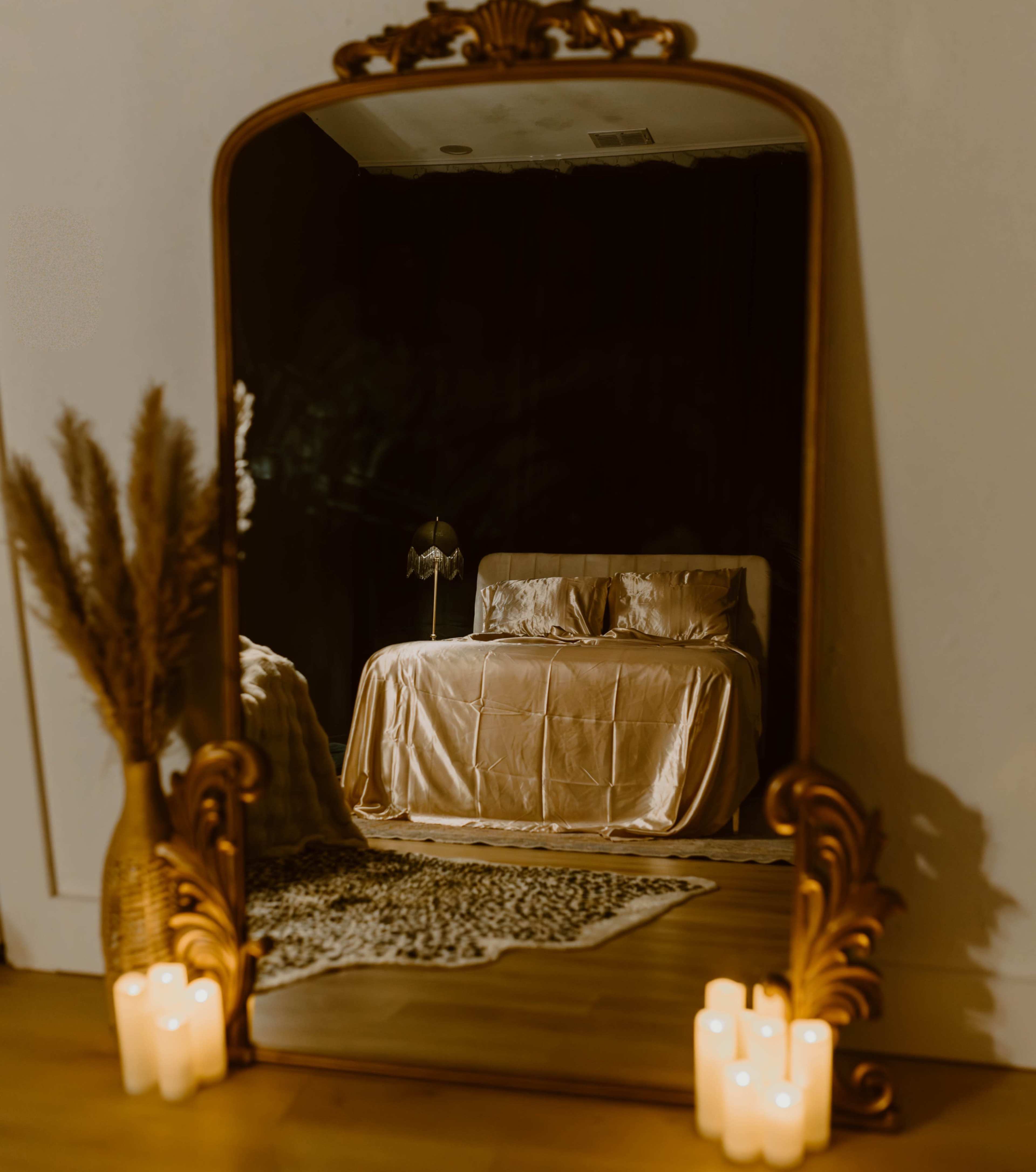 A large, ornate mirror reflects a softly lit bedroom featuring a bed with satin bedding and a decorative lamp beside it, surrounded by candles and a plant.