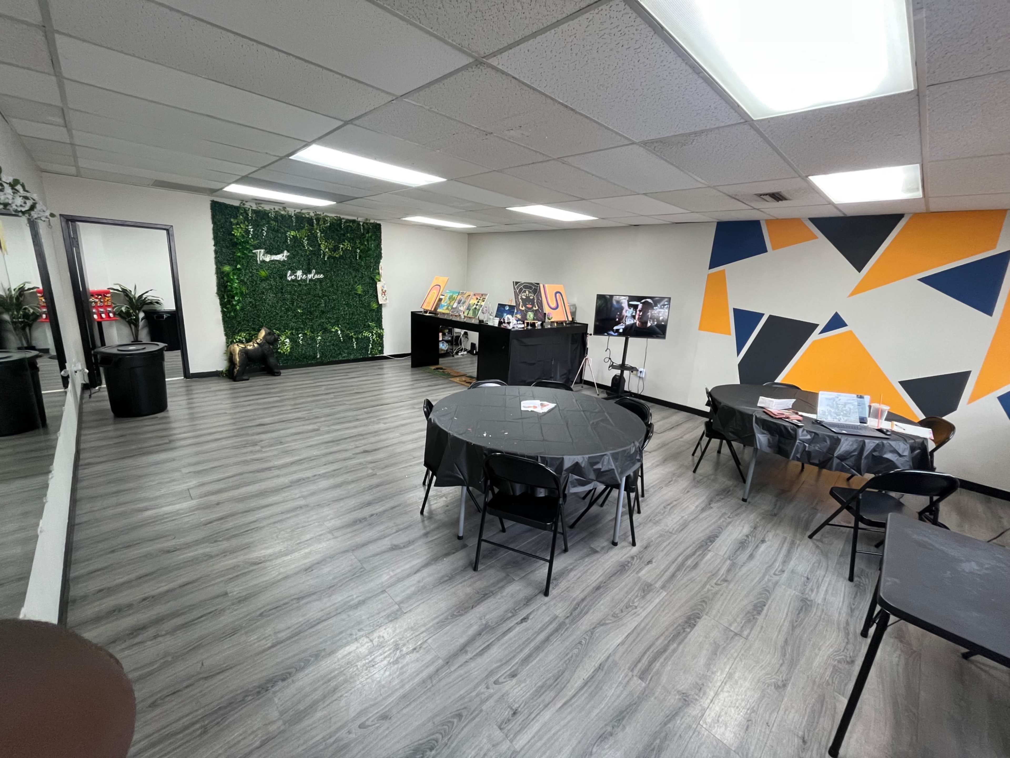 The room features several round tables with black tablecloths, a display wall with greenery, and colorful geometric shapes painted on one wall.