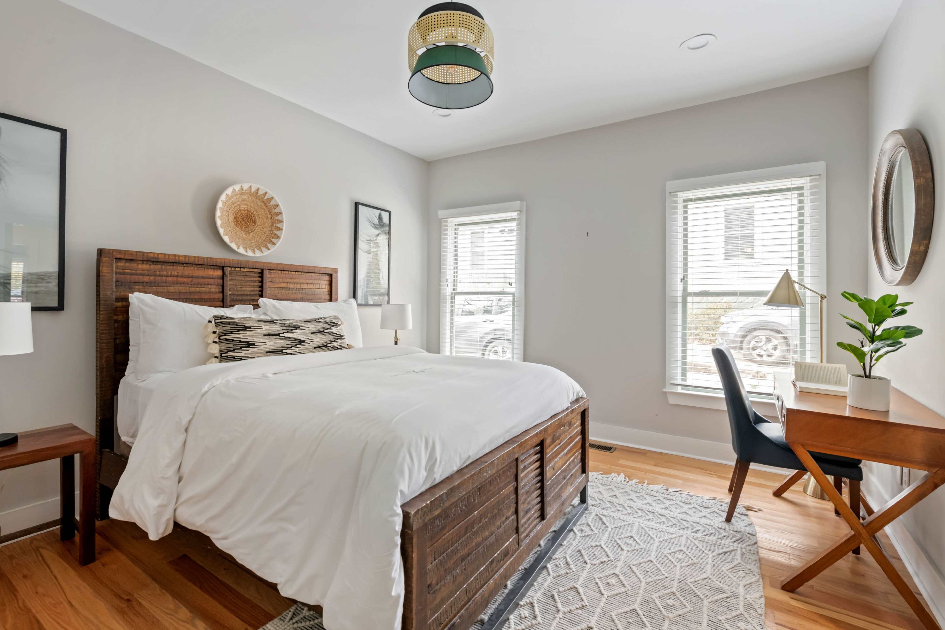 The image shows a well-lit bedroom featuring a large bed, a small desk with a chair, and two windows with natural light.