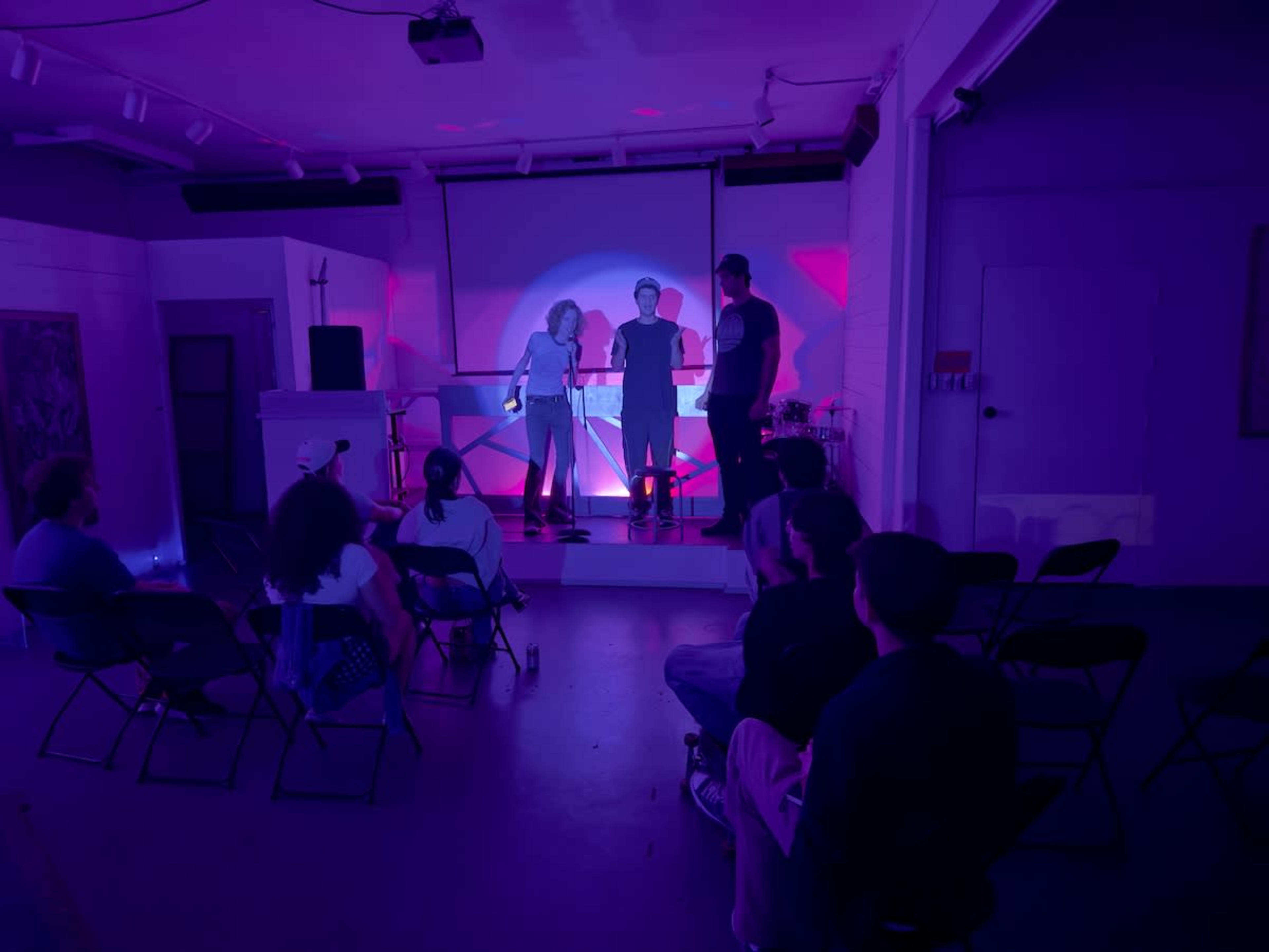 A small audience sits on folding chairs while three performers engage on a brightly lit stage with purple lighting.