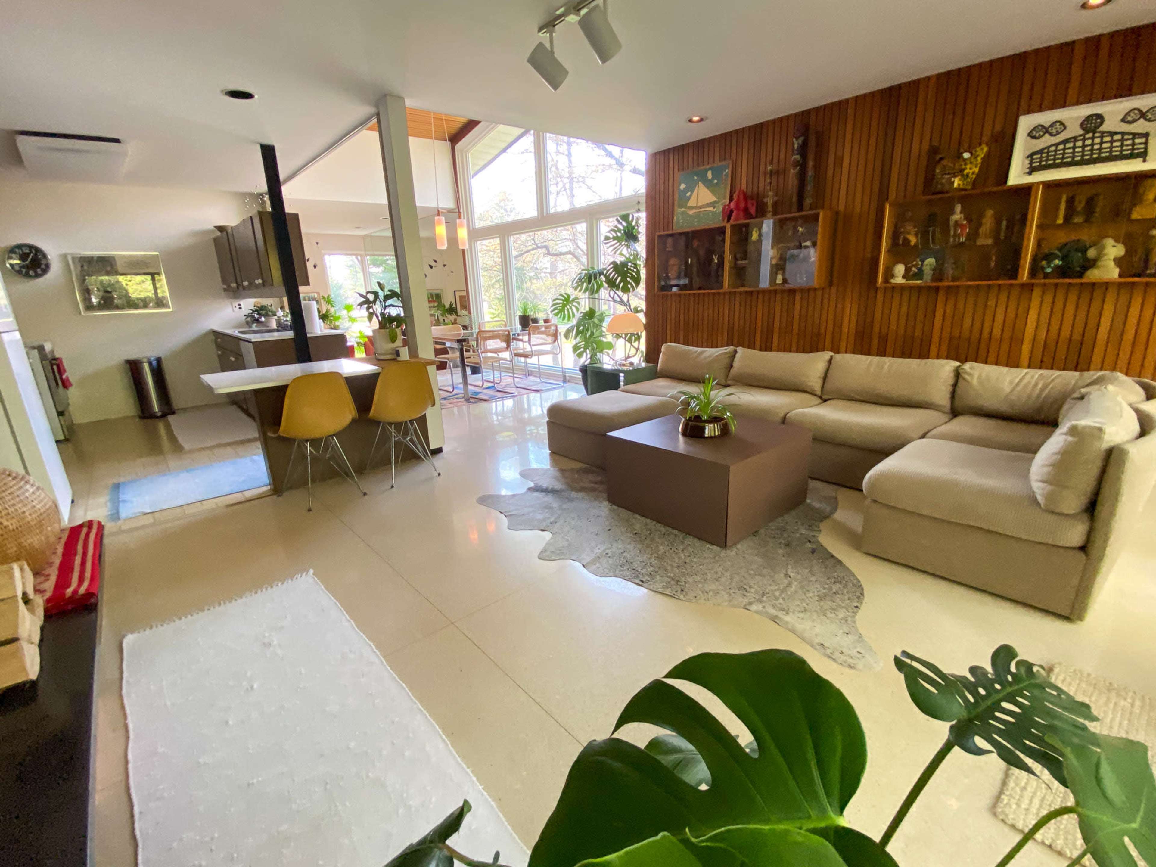 The image shows a modern living room with a large sectional sofa, a coffee table, and an open layout connecting to a dining area filled with plants.