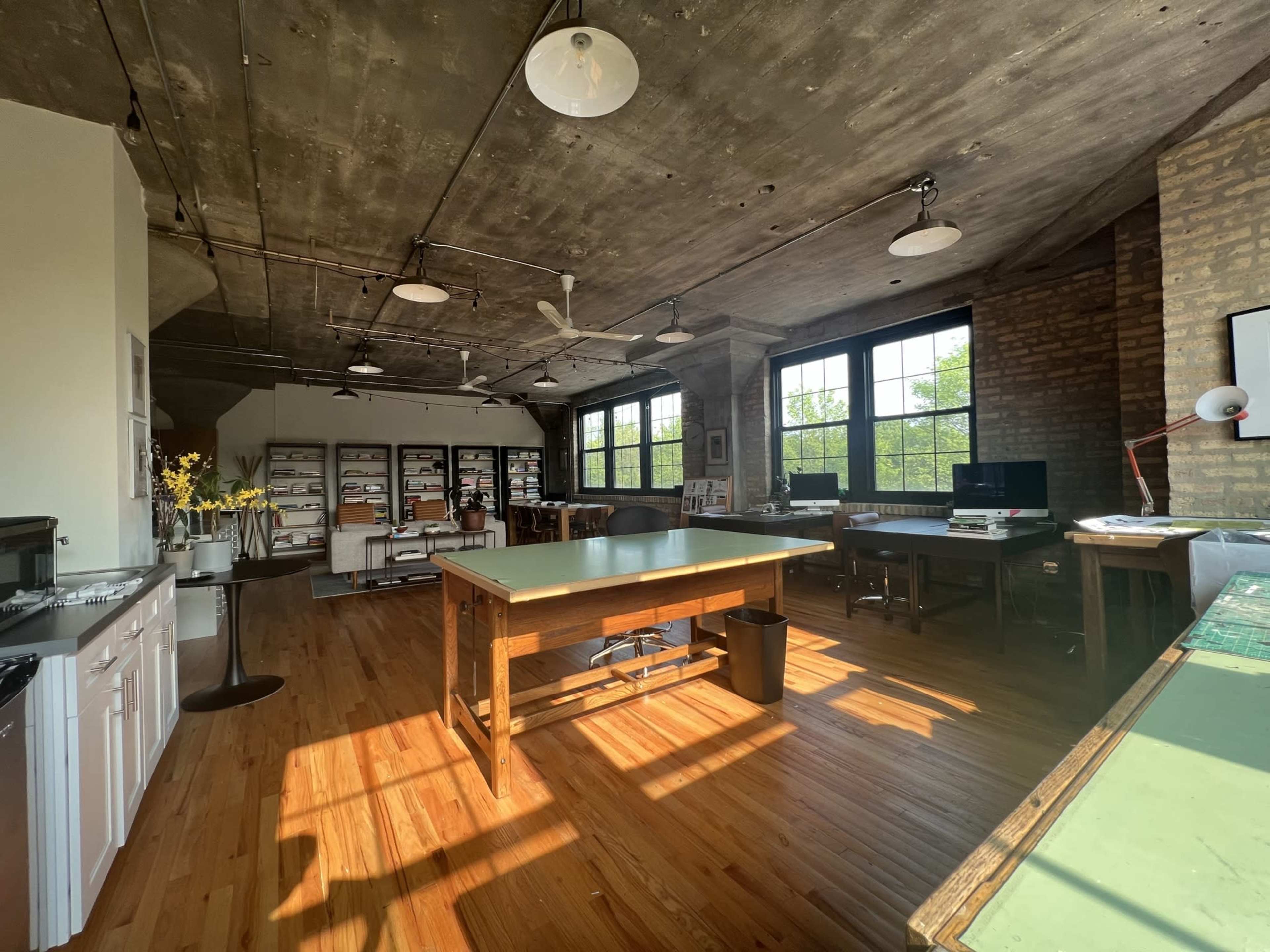 An open office space with wooden floors, large windows, and a central worktable, surrounded by various workstations and shelves.