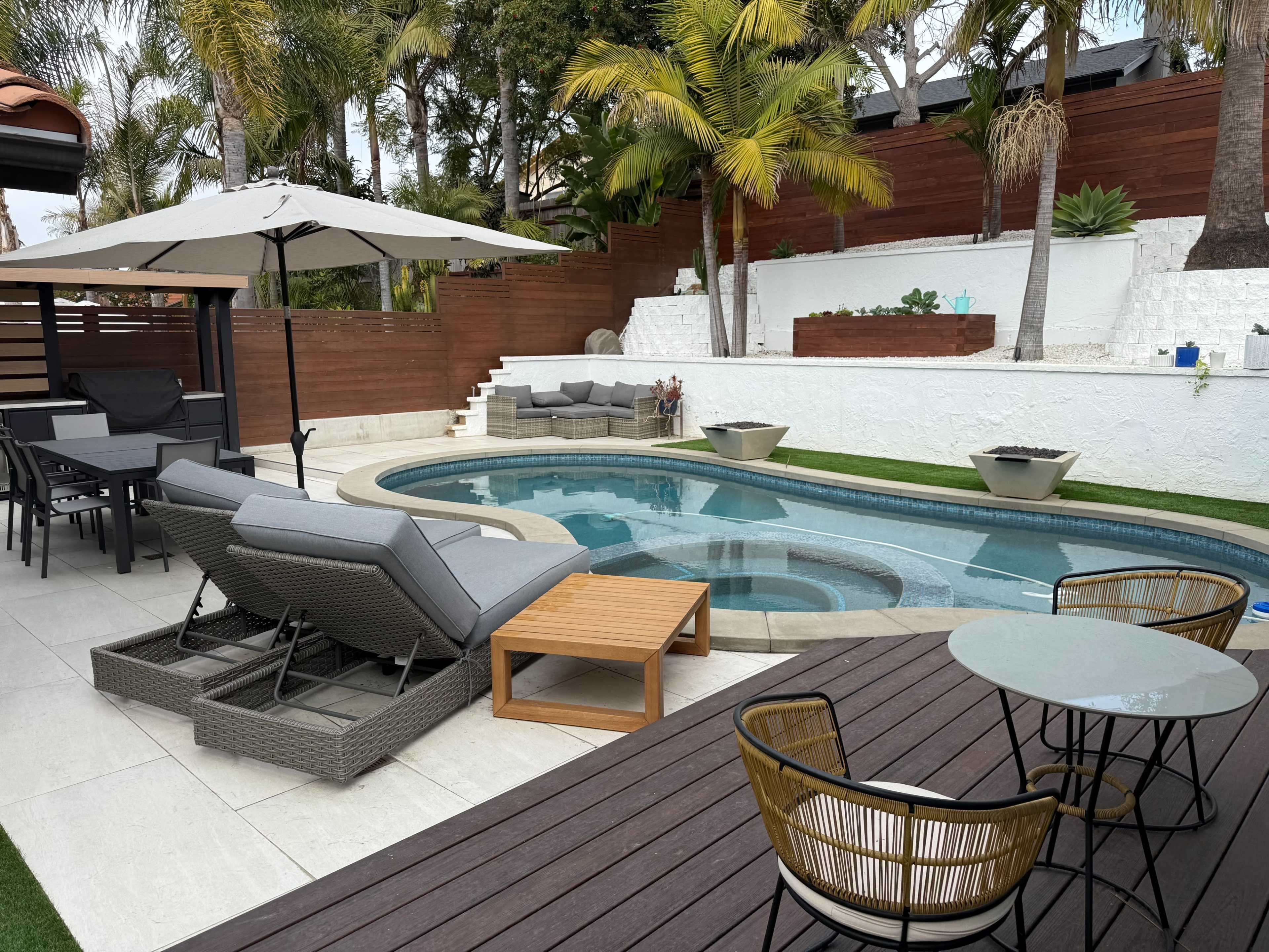 The image depicts a modern backyard oasis featuring a swimming pool surrounded by lounge chairs, a dining area, and tropical landscaping.