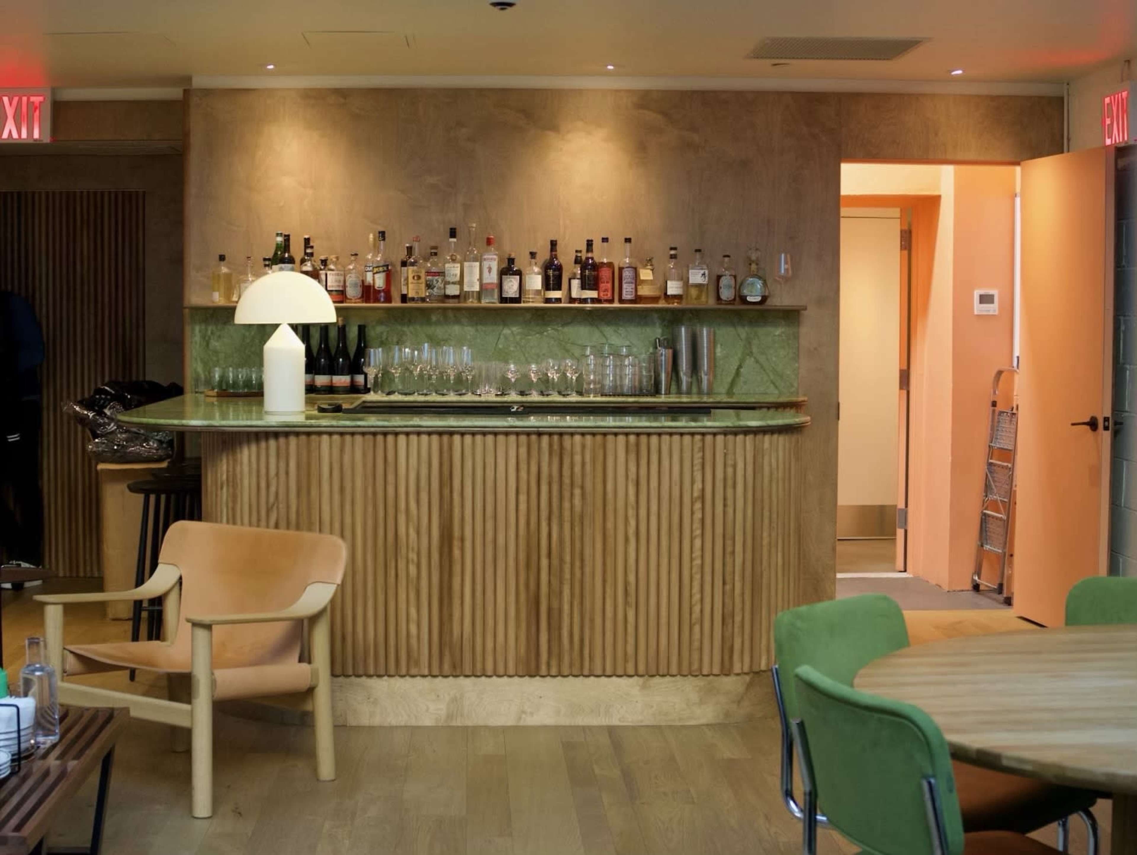 A wooden bar with a green stone countertop holds various bottles and glasses, and a doorway leads to an orange-lit room beyond.