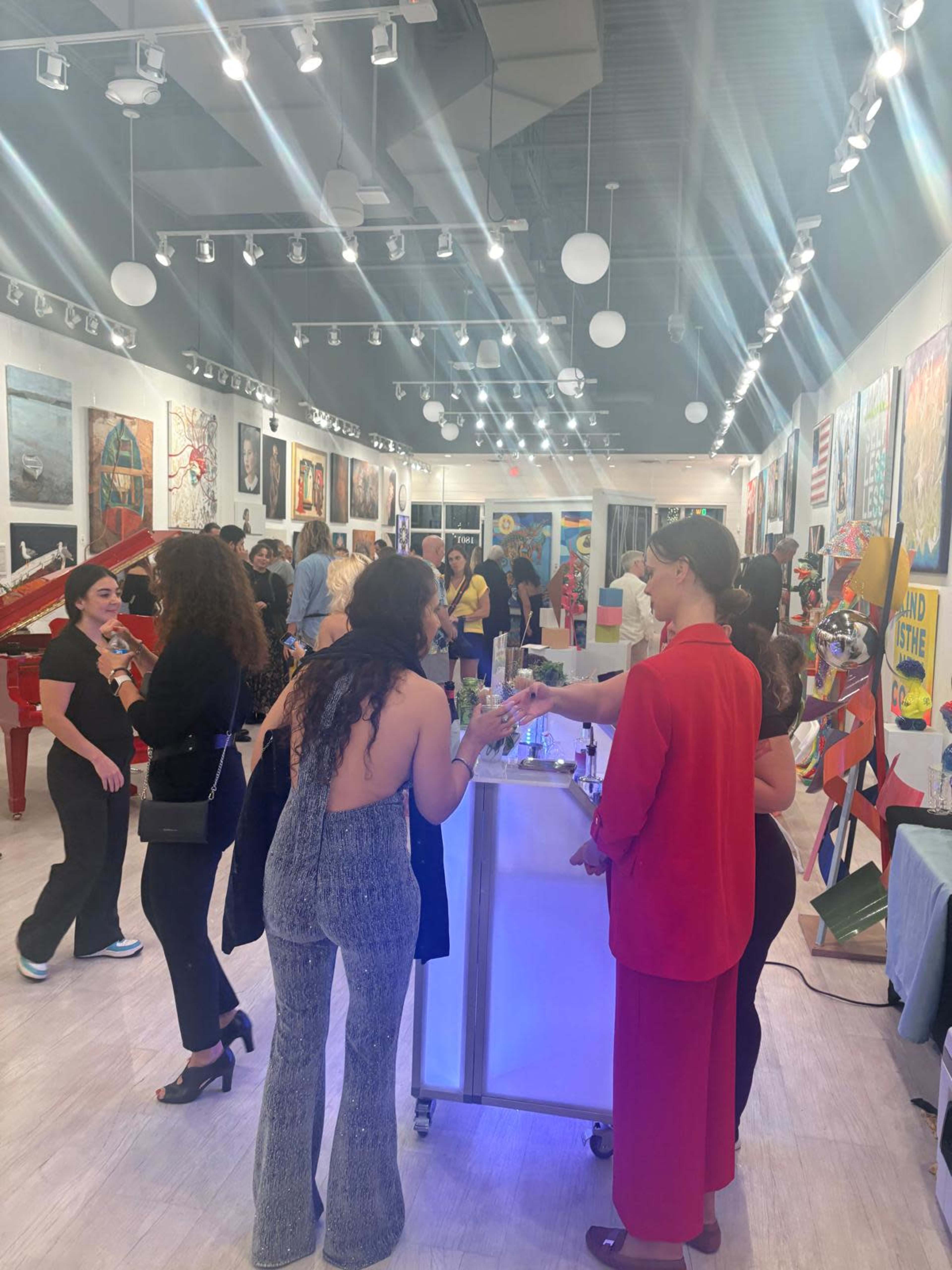 A lively art gallery event showcases attendees mingling, with various artworks displayed on the walls and a red piano in the center.