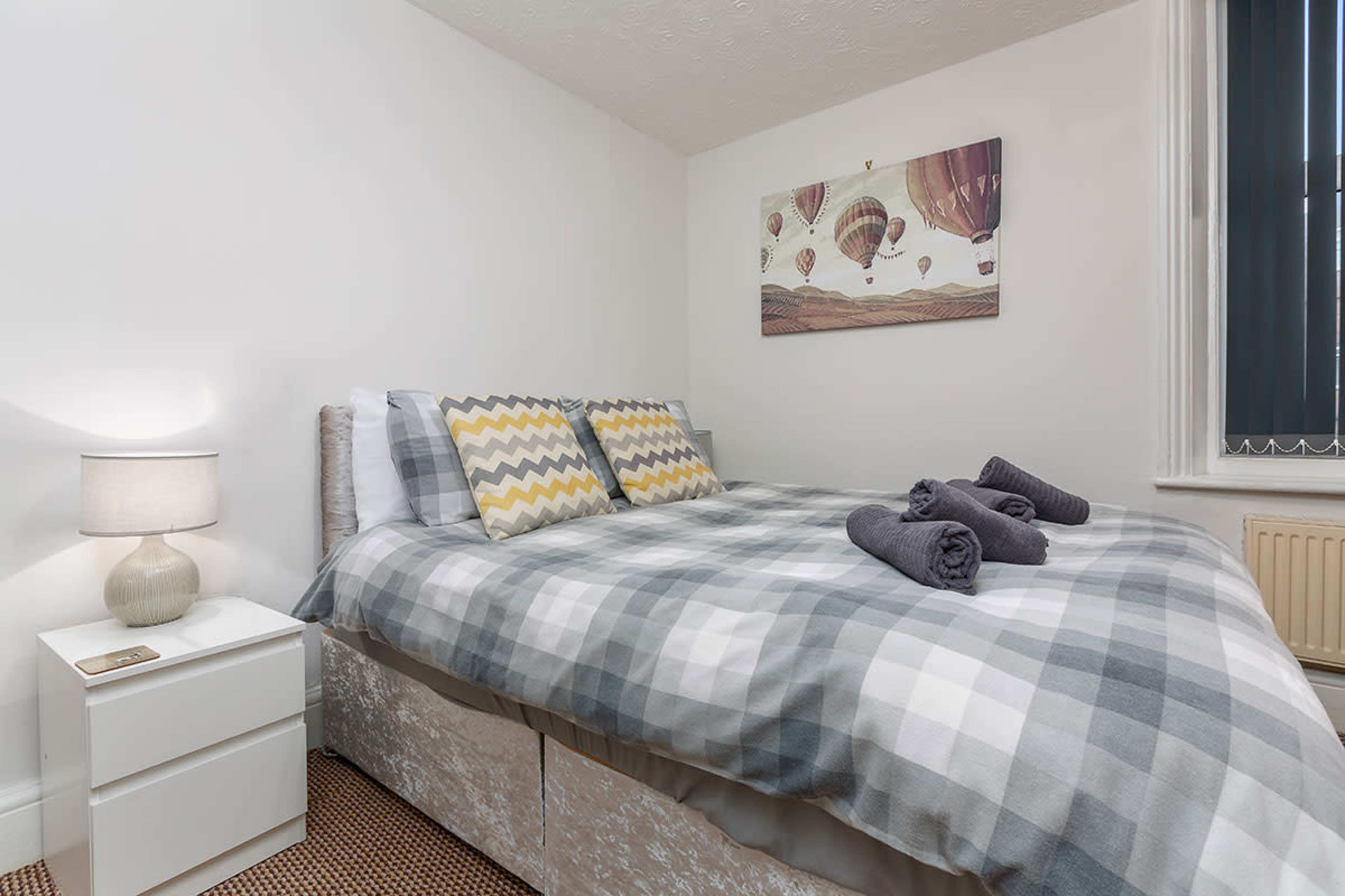 A neatly arranged bedroom features a double bed with gray and white checkered bedding, two decorative pillows, and rolled towels, alongside a bedside table with a lamp.