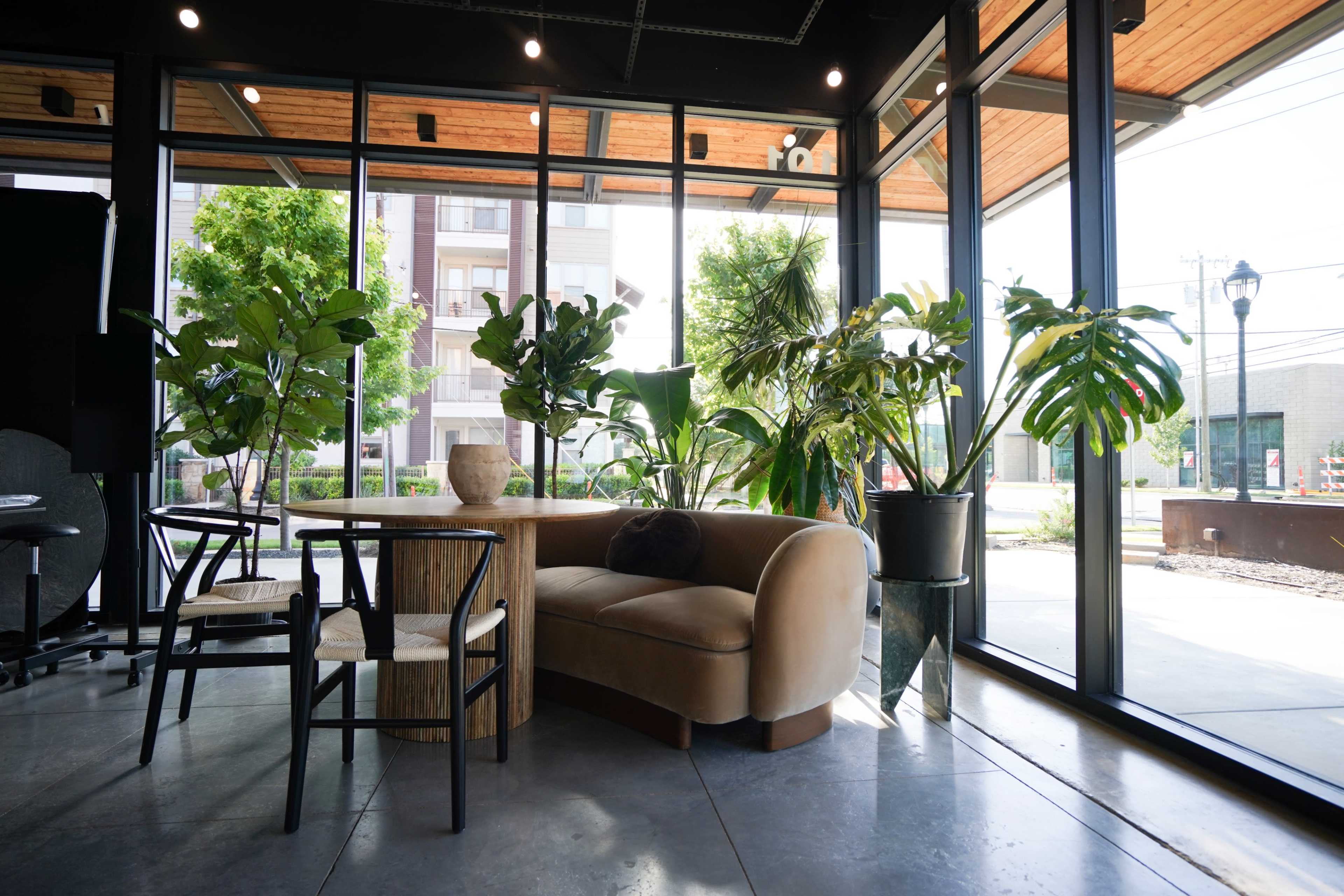 A cozy seating area is set within a glass-walled space, featuring a curved sofa, a wooden table, and various potted plants.